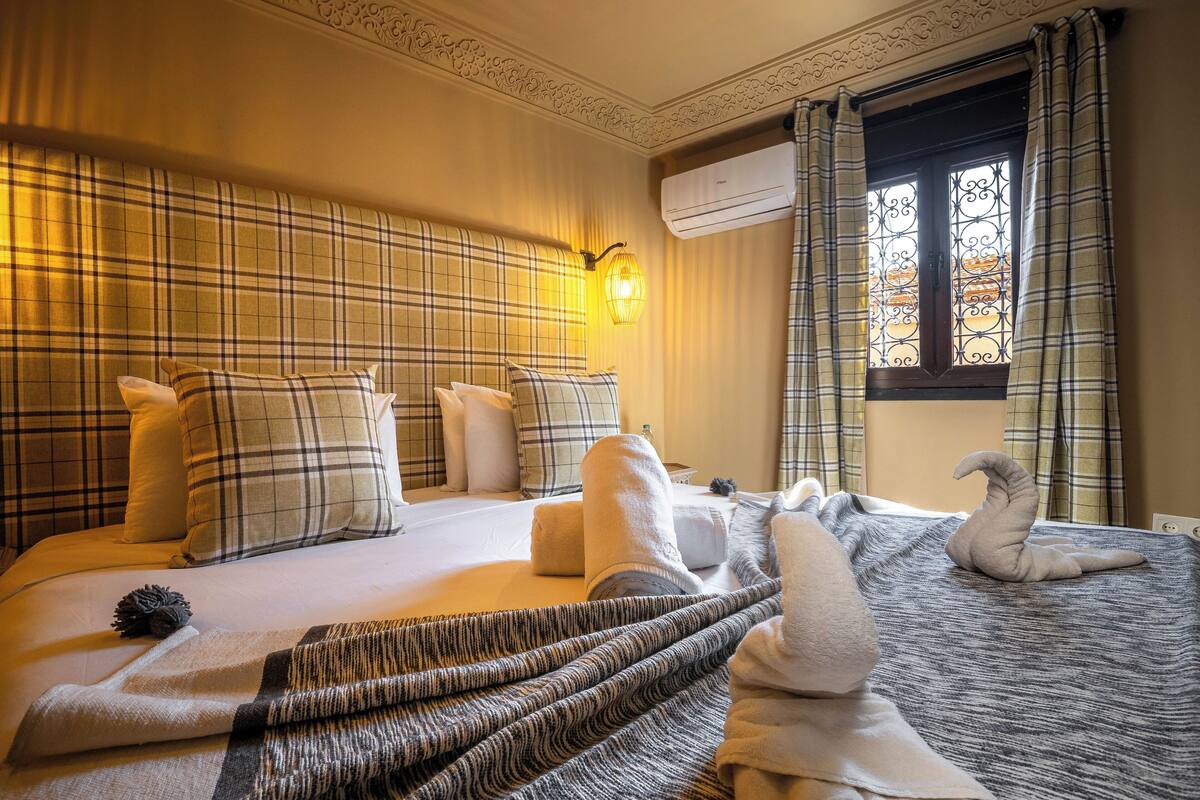 A hotel room with a bed, pillows, a blanket, a wall-mounted light, an air conditioning unit, and a window with curtains.