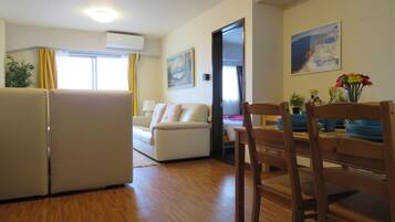 A compact living space with a sofa, dining table, and chairs. There is a window with curtains, a painting on the wall, and an air conditioning unit.