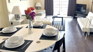 Een eethoek met een ronde tafel voor vier personen, een vaas met bloemen en uitzicht op een woonkamer met een bank en een televisie.