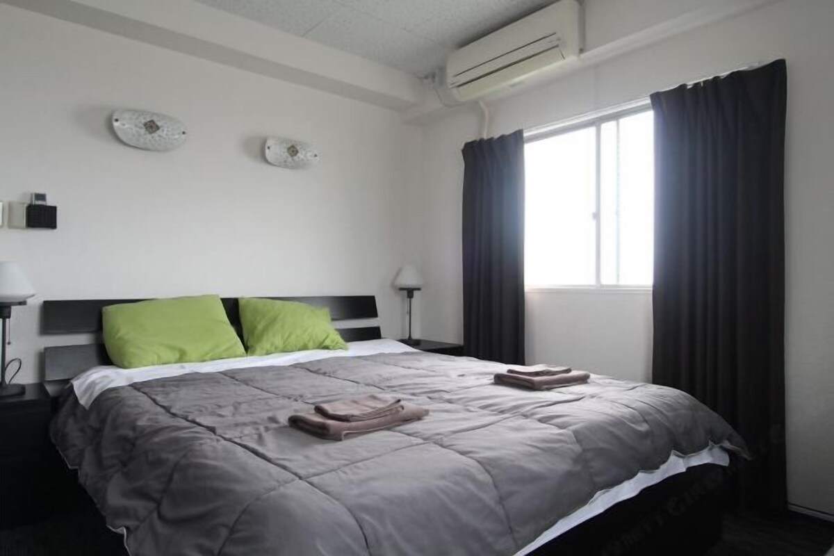 A hotel room with a bed, two pillows, a nightstand, a lamp, a window with curtains, and an air conditioning unit.