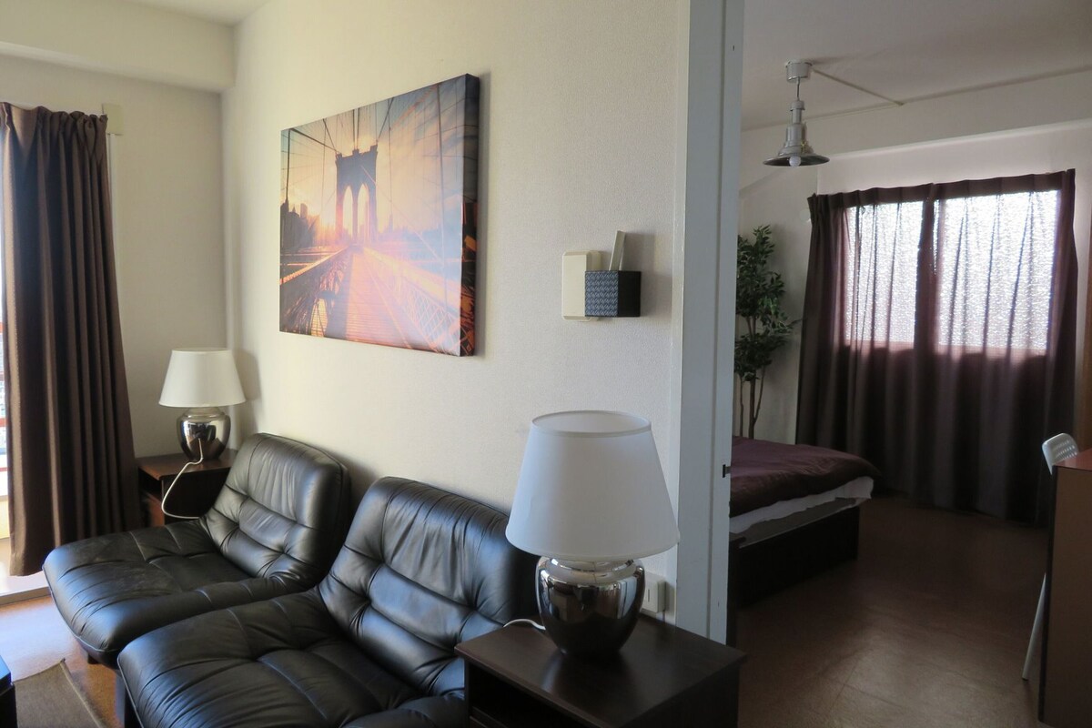 A room with a sofa, two lamps, a painting, a bed, and a window with curtains.