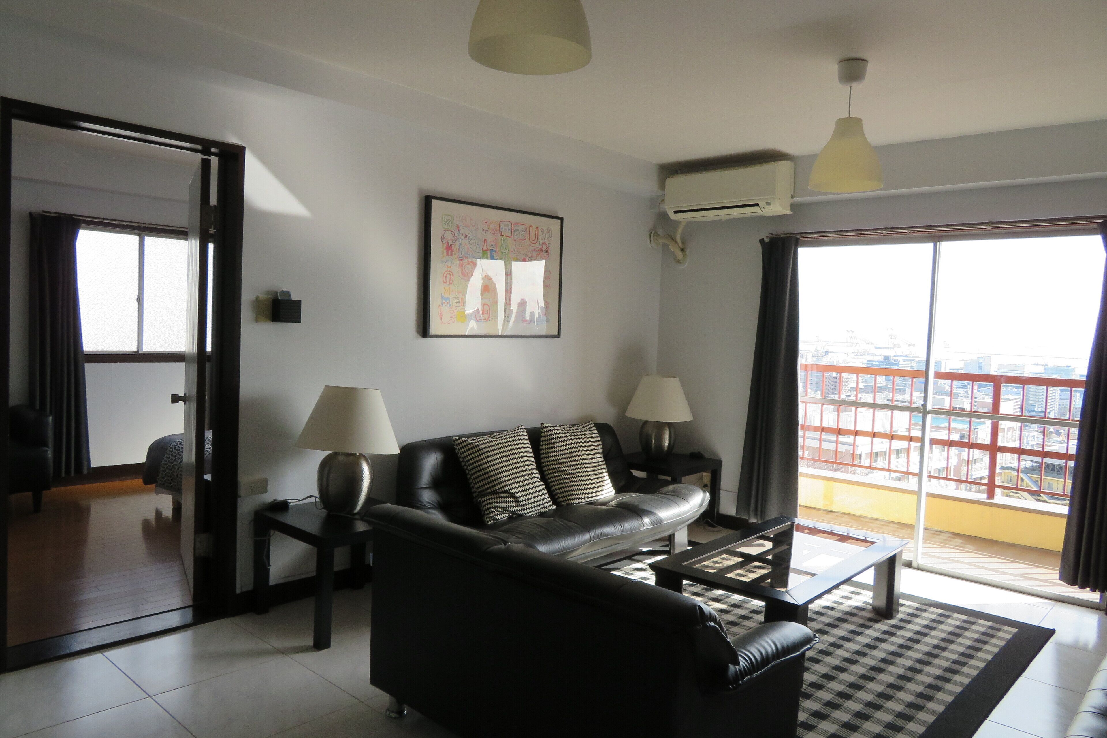 A modern living room with a black leather sofa, two side tables with lamps, a framed artwork on the wall, and a large window with a city view.