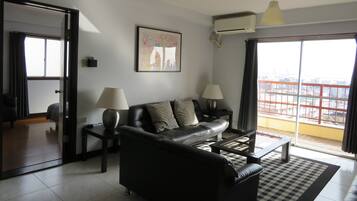 A modern living room with a black leather sofa, two side tables with lamps, a framed artwork on the wall, and a large window with a city view.