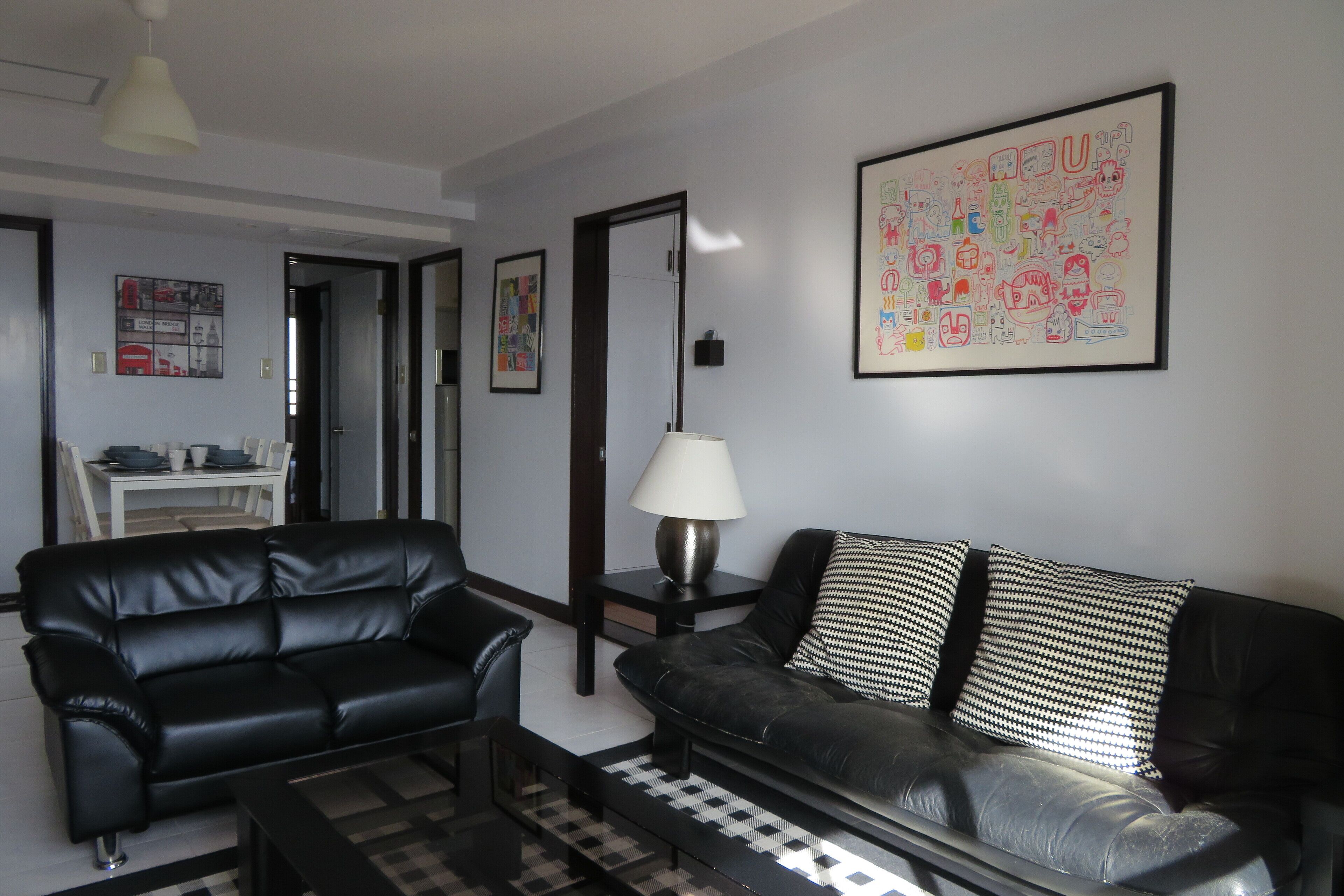 A modern living room with a black leather sofa, a coffee table, and a lamp. There is a framed picture on the wall and a door leading to another room.