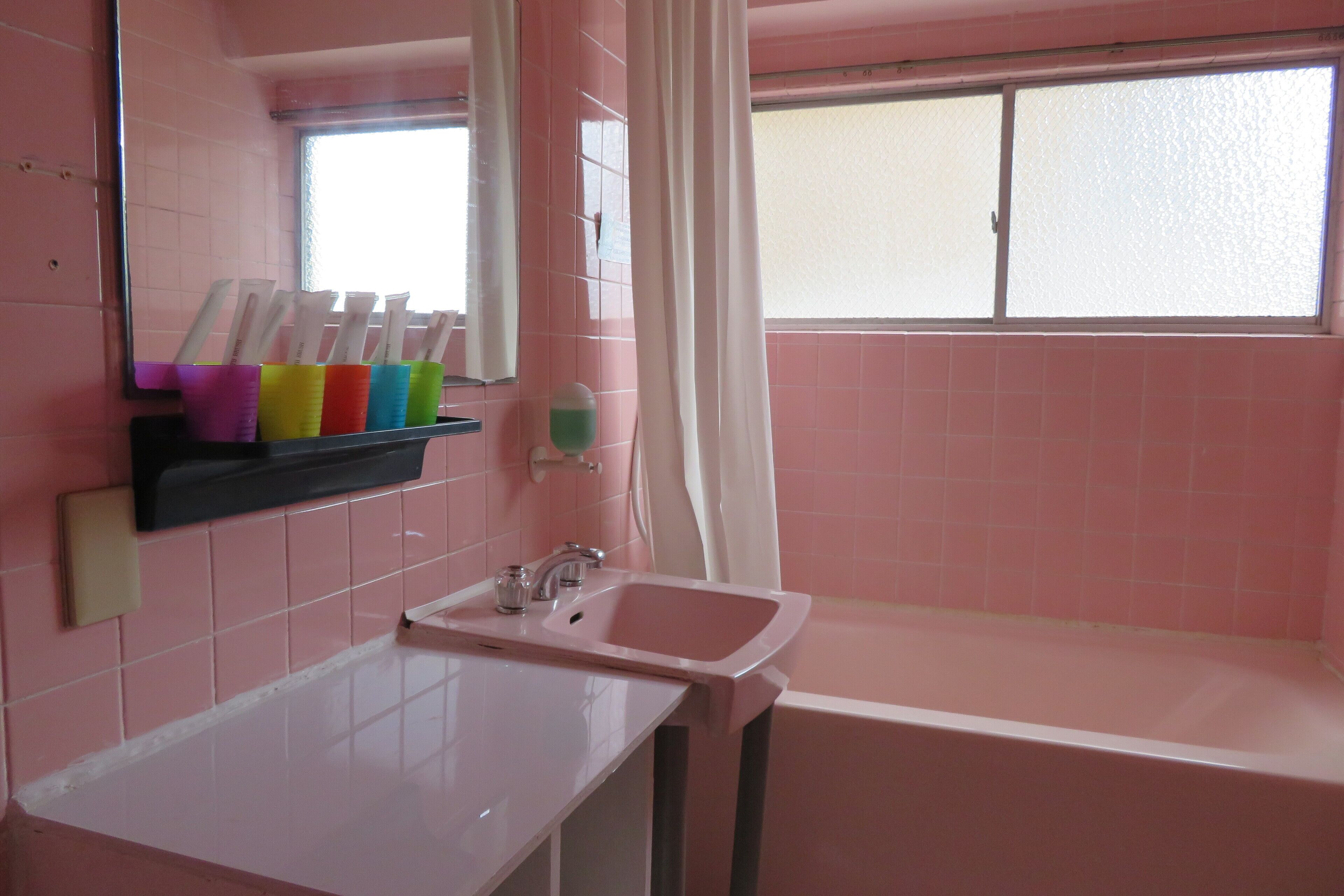 Ein pinkfarbenes Badezimmer mit Waschbecken, Spiegel und einem Fenster mit Vorhang.