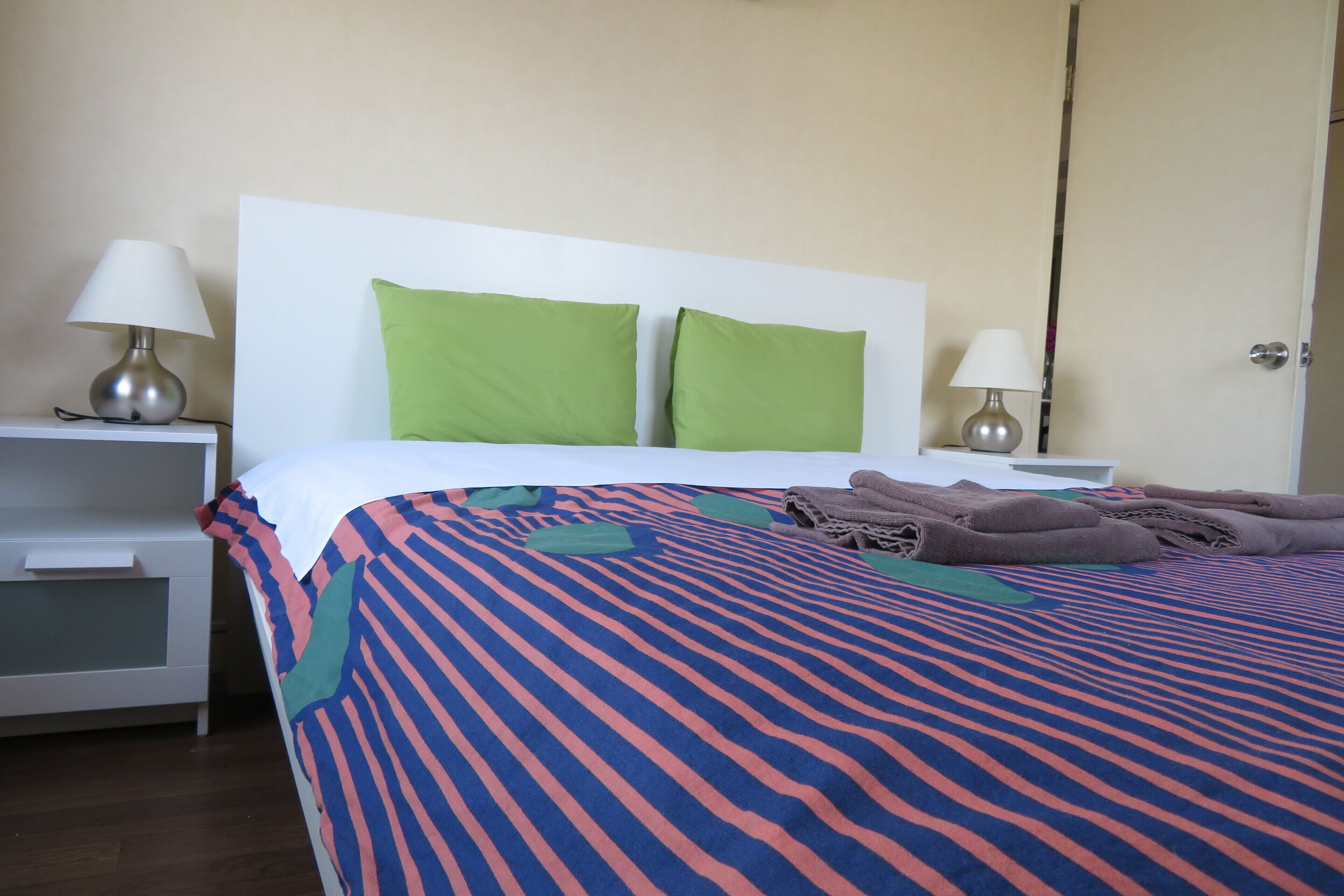 Una cama bien tendida con ropa de cama a rayas, dos almohadas verdes y toallas dobladas sobre un cabecero blanco.