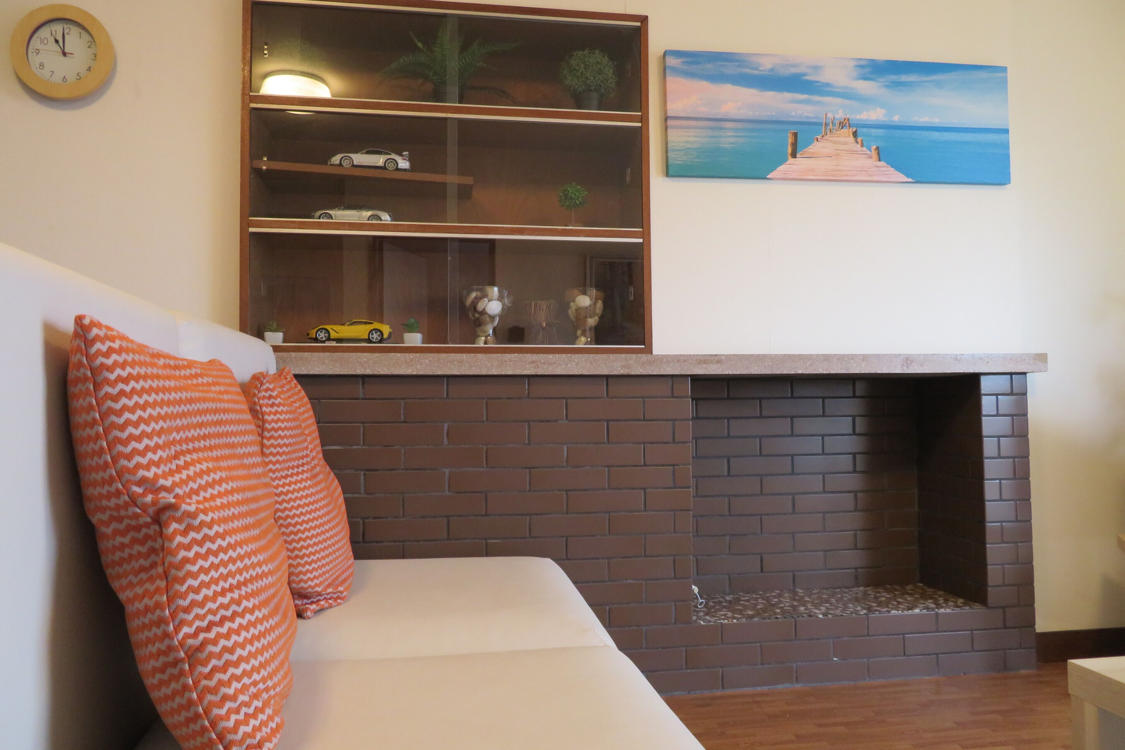 A living room with a brick fireplace, a white sofa with orange pillows, a shelf with decorative items, and a painting of a pier.