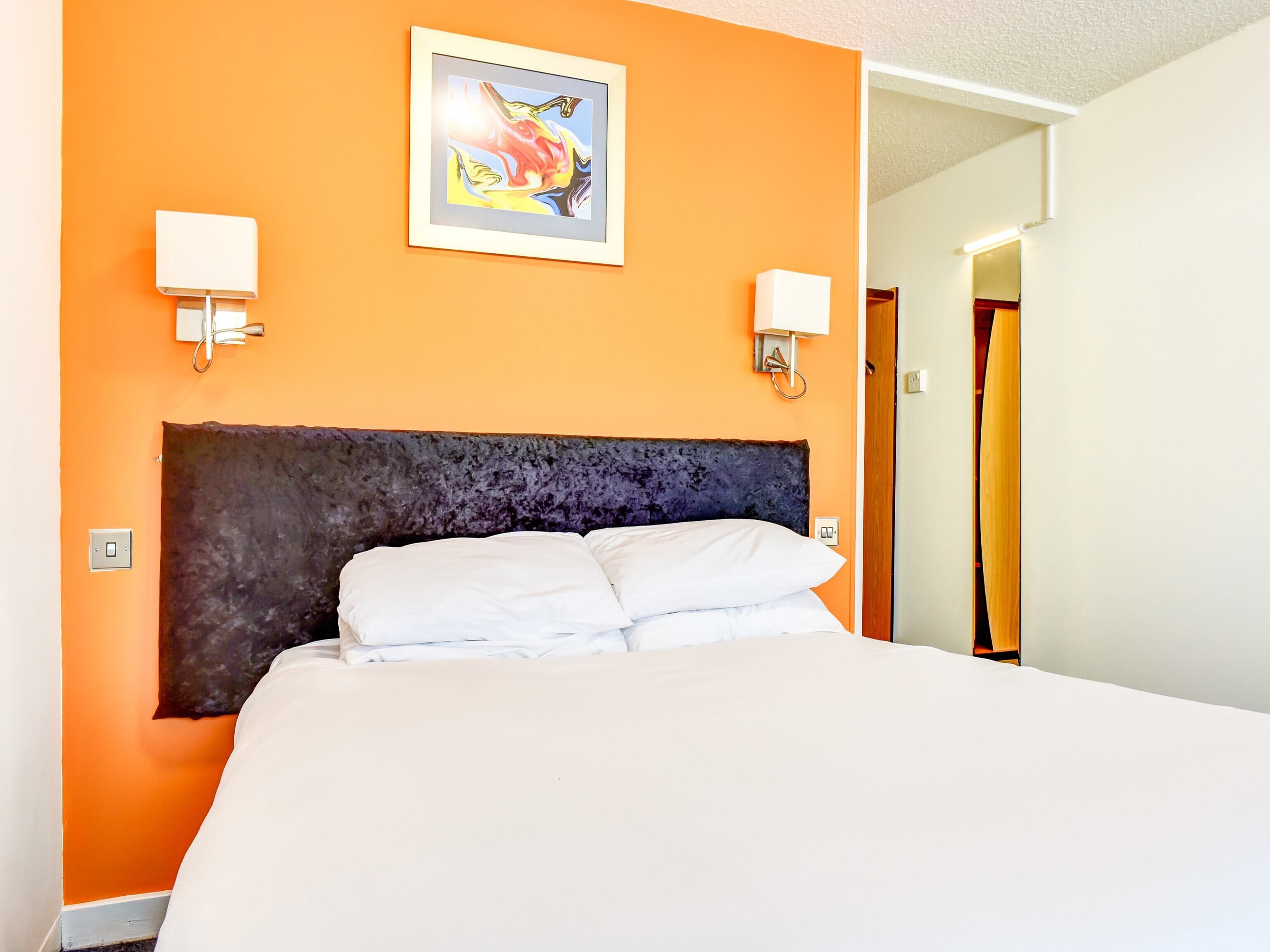 Hotelkamer met een oranje wand, een donker hoofdeinde, twee bedlampen en een ingelijste afbeelding boven het bed.