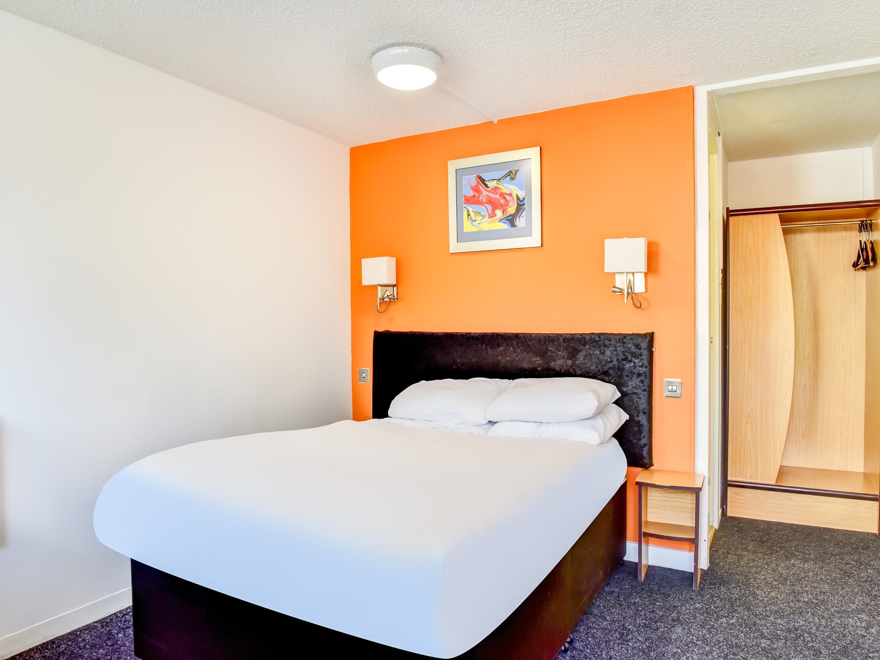 A hotel room with a large bed, an orange accent wall, a painting, two wall sconces, and a wooden closet.