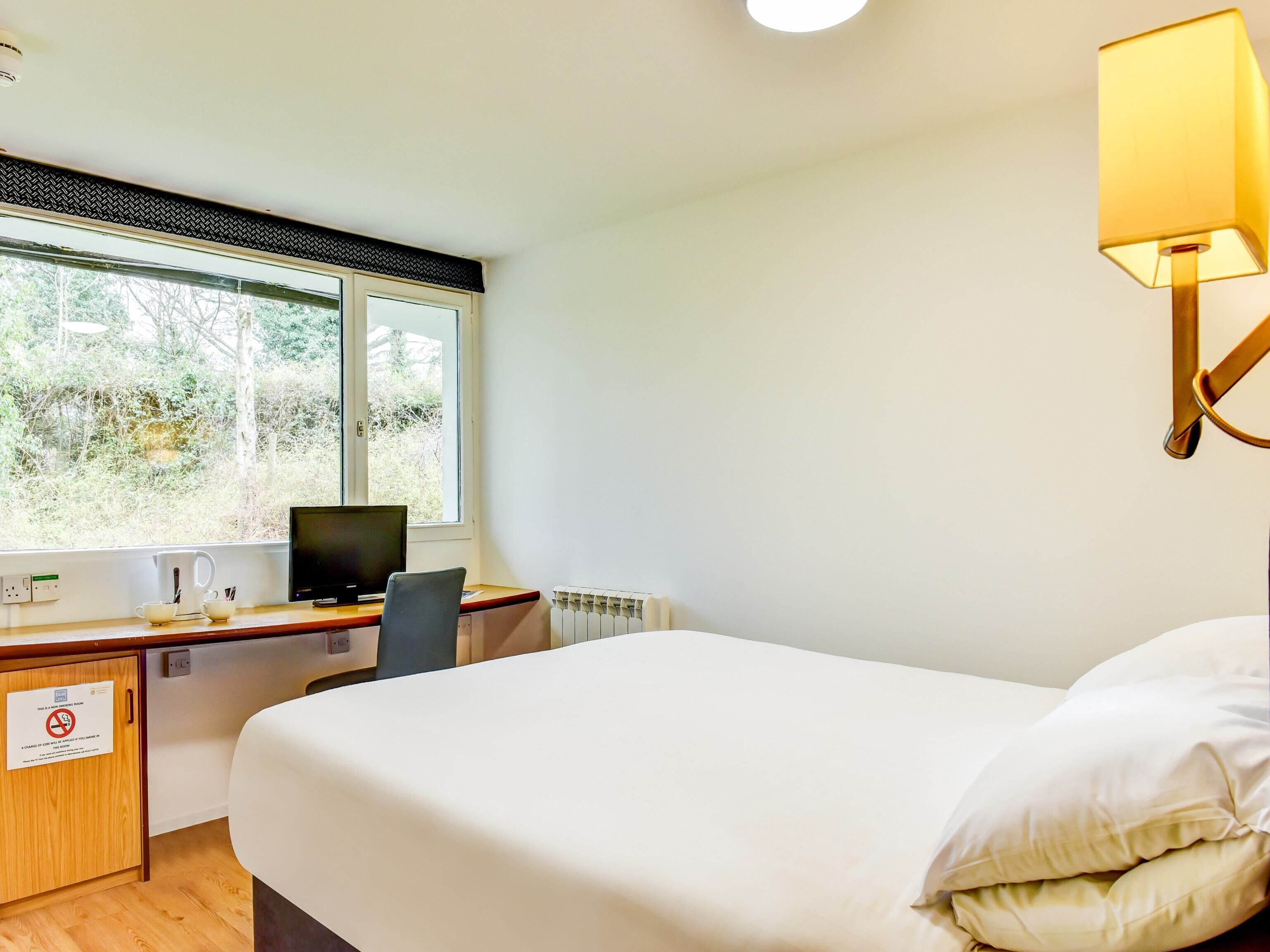 Ein Hotelzimmer mit Bett, Schreibtisch, Stuhl, Computer und einem Fenster mit Blick auf Bäume.