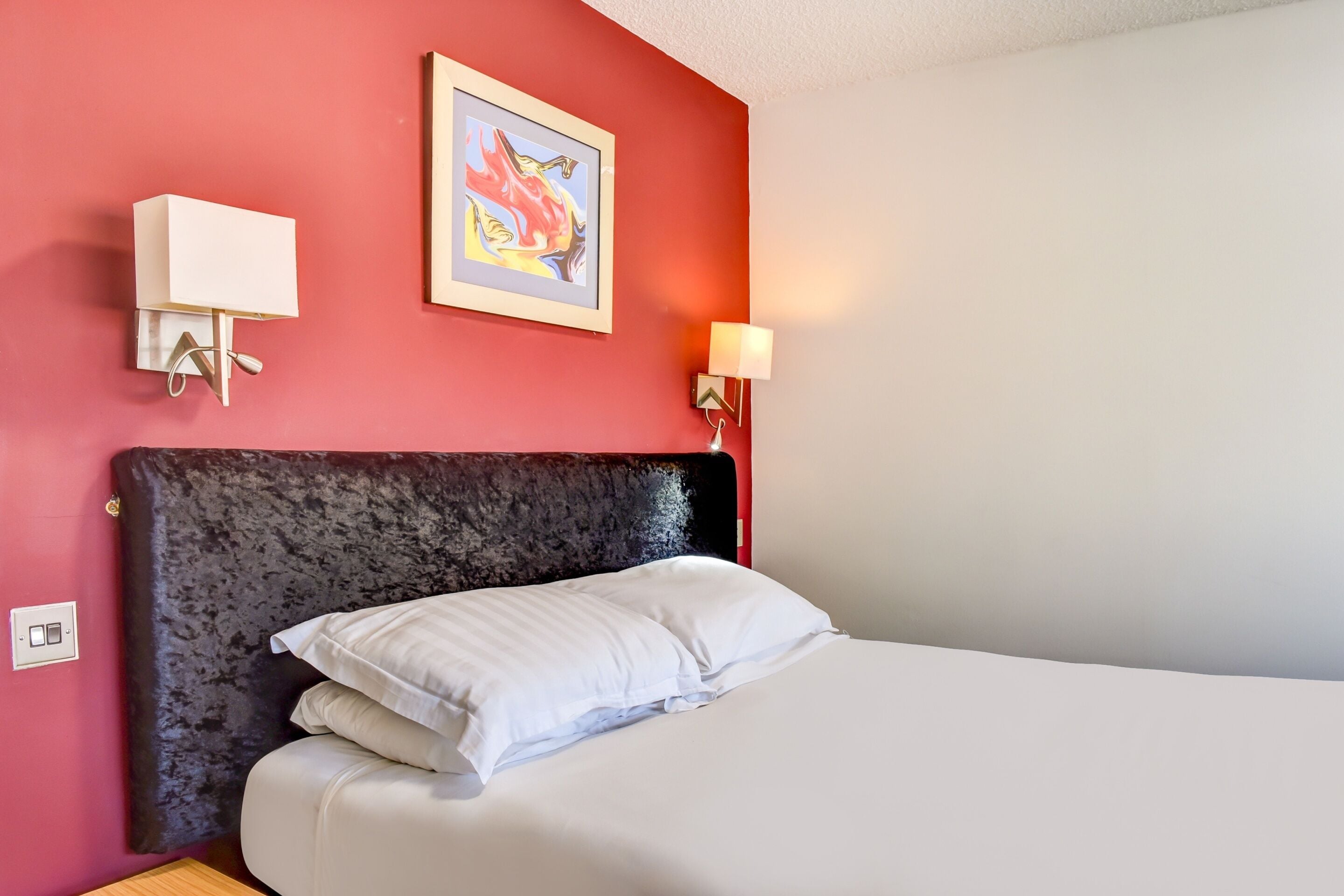 A hotel room with a red wall, a framed artwork, a bed with white bedding, and a wall-mounted light fixture.