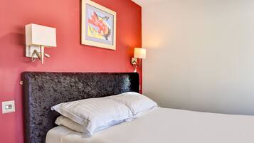A hotel room with a red wall, a framed artwork, a bed with white bedding, and a wall-mounted light fixture.