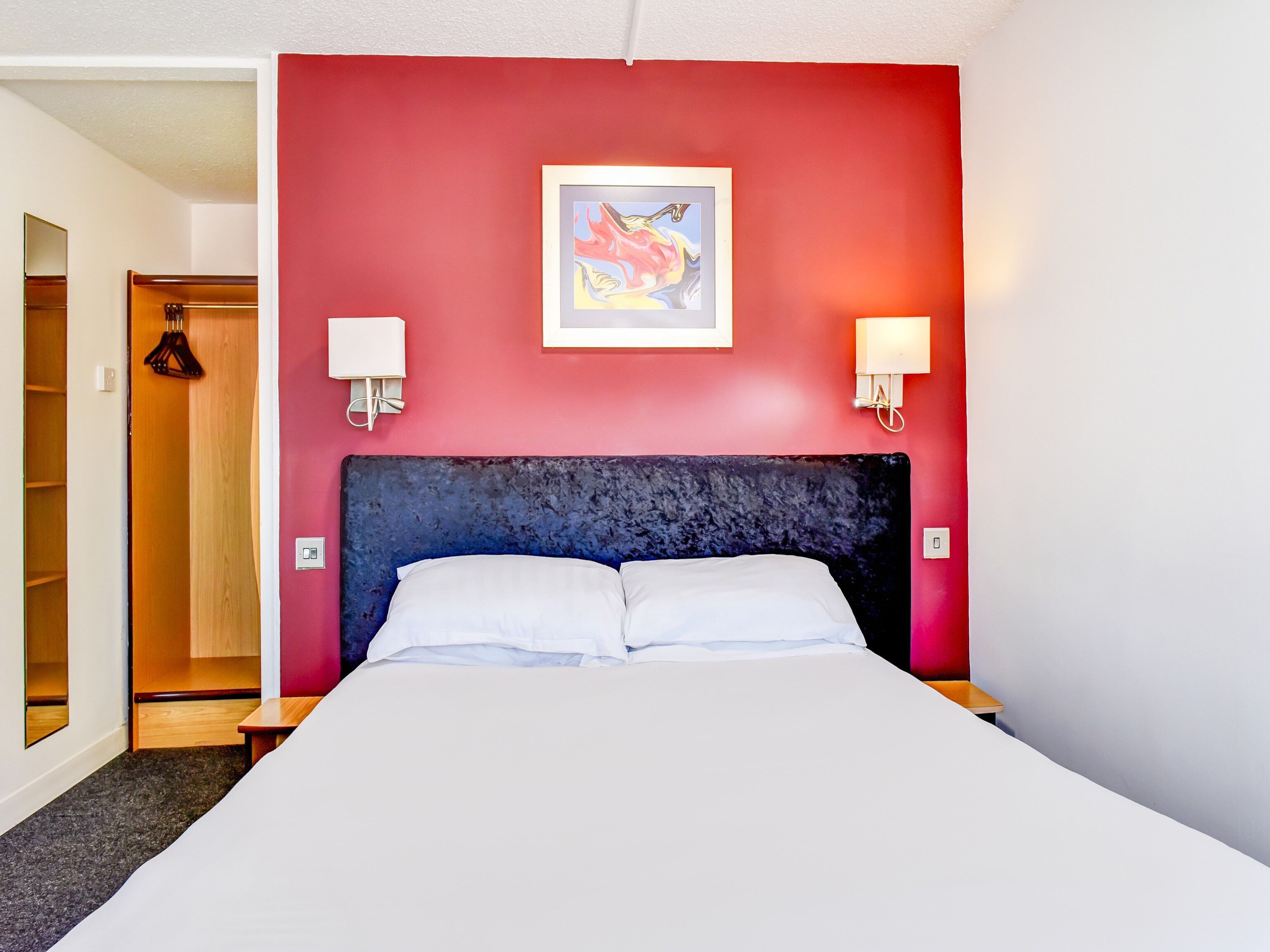 A hotel room with a red accent wall, a large bed with white linens, two bedside lamps, and a framed artwork above the bed.