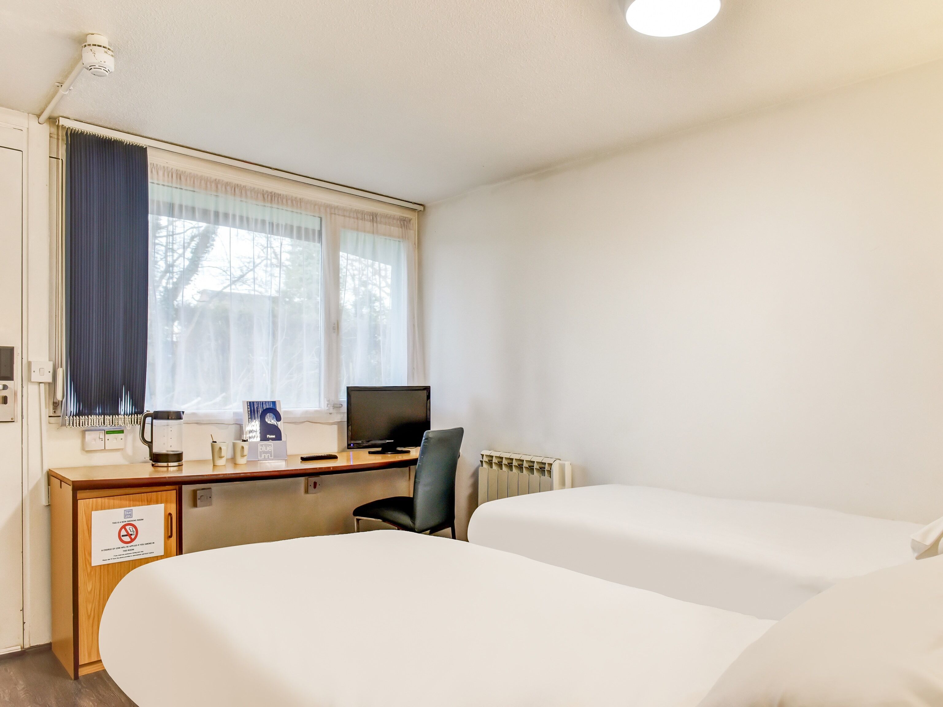 Habitación de hotel con escritorio, computadora, silla, dos camas, ventana con cortinas y radiador.