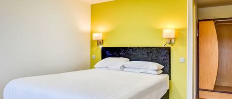 A neatly made bed in a room with a yellow accent wall, a wooden wardrobe, and two wall-mounted lamps.