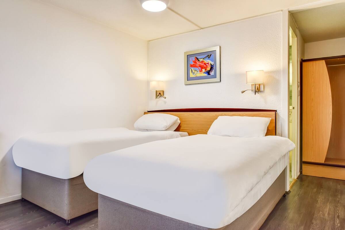 A hotel room with two beds, a wooden headboard, a painting on the wall, and two wall-mounted lamps.