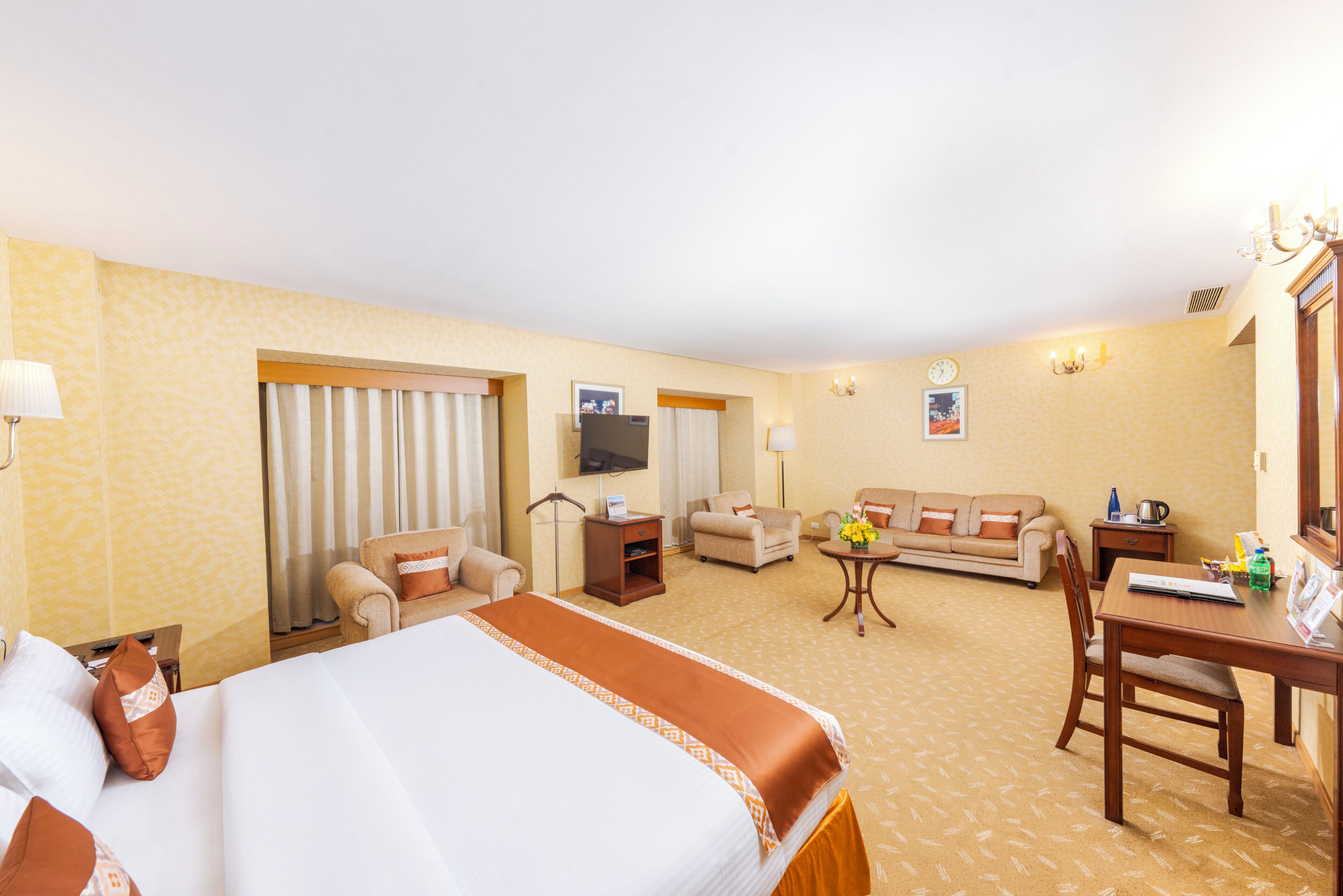Quarto de hotel com cama, duas poltronas, um sofá, televisão, escrivaninha e uma pequena mesa com flores.
