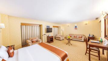 Quarto de hotel com cama, duas poltronas, um sofá, televisão, escrivaninha e uma pequena mesa com flores.