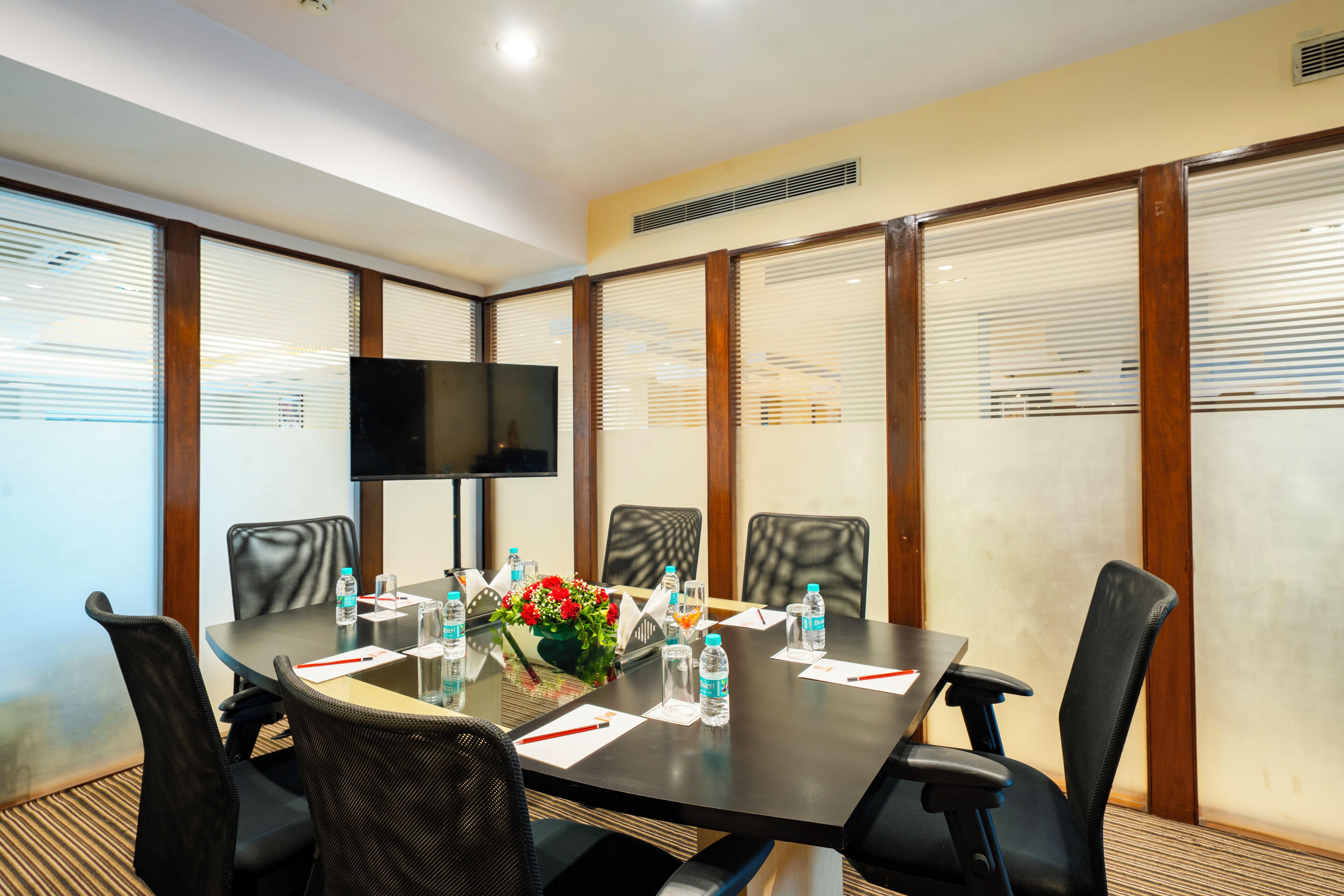 A conference room with a glass table, black chairs, a TV screen, and a floral arrangement.