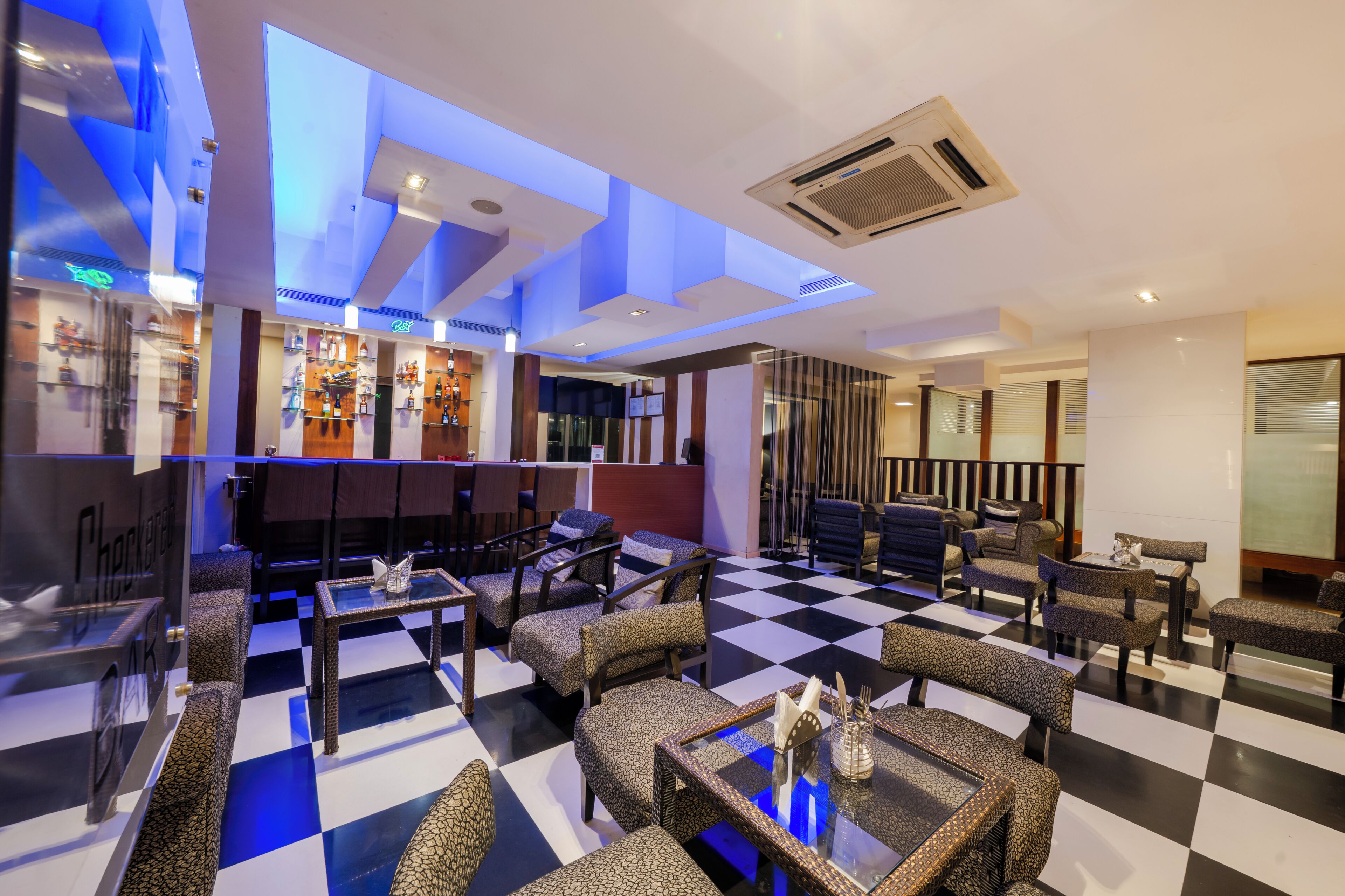 Un vestíbulo de hotel con piso a cuadros, zonas de estar, un bar con estantes llenos de botellas e iluminación empotrada en el techo.