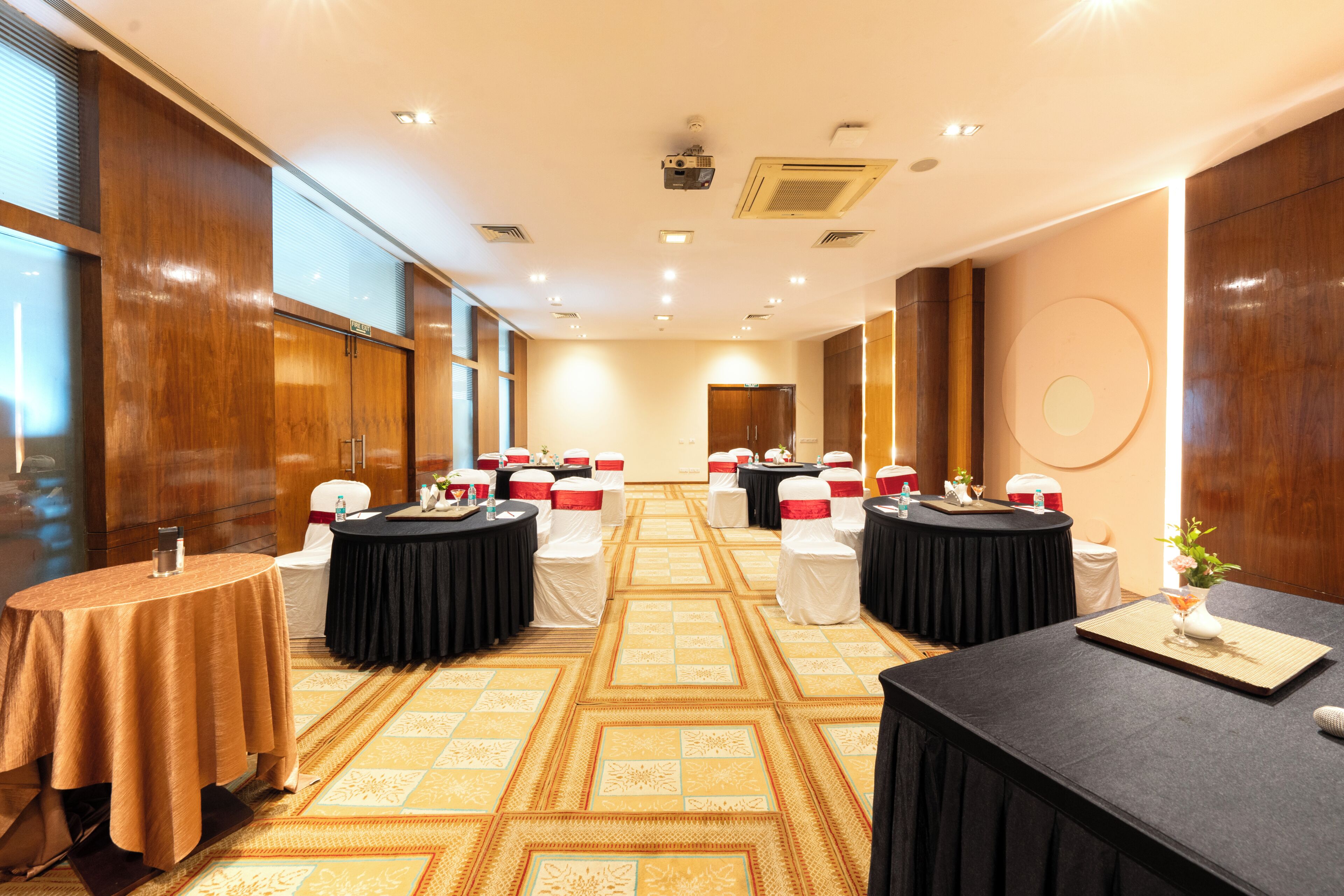 A hotel conference room with multiple round tables, chairs, and a carpeted floor.