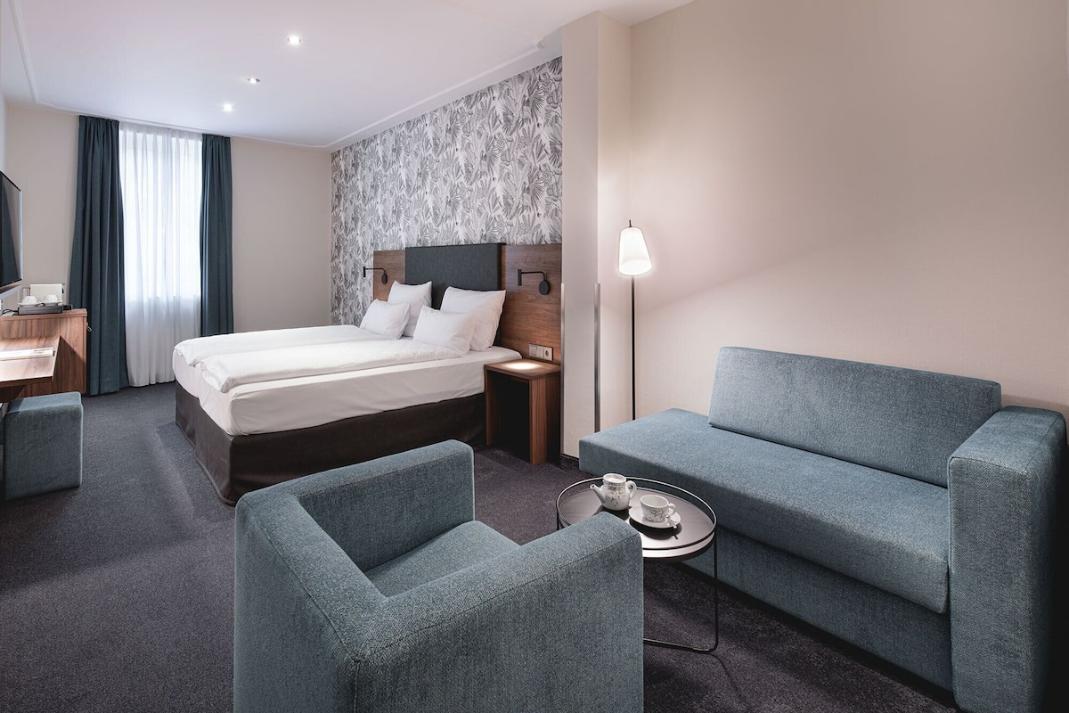 A hotel room with a bed, two armchairs, a small table with a teapot, a desk, and a TV.