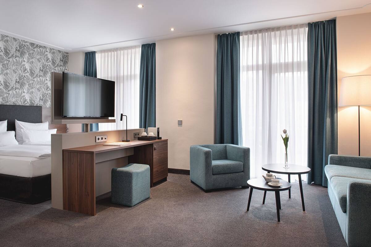 A modern hotel room with a bed, desk, television, two armchairs, a coffee table, and a vase with a flower.