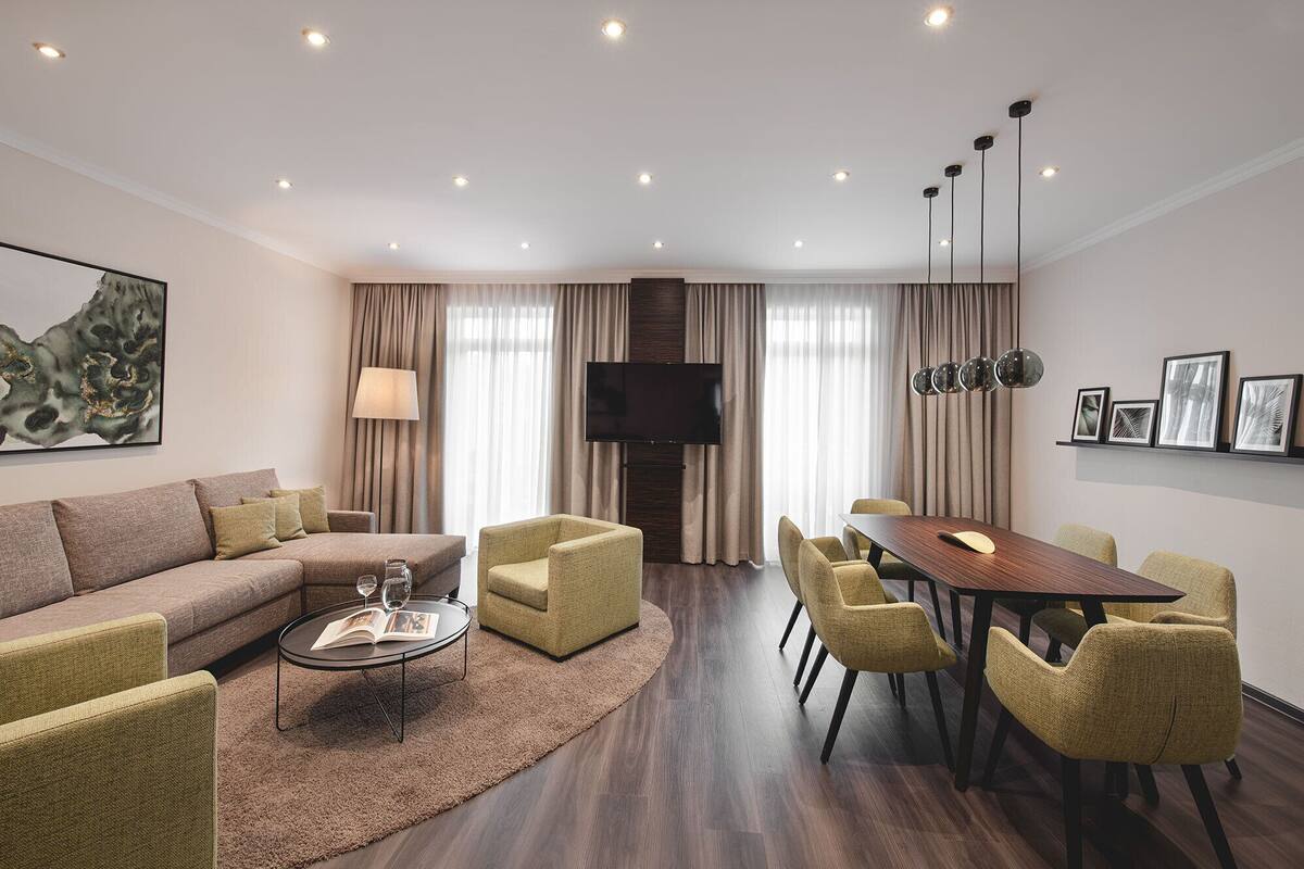 A modern living room with a sofa, armchairs, a coffee table, and a dining table with chairs. A flat-screen TV is mounted on the wall, and there are ceiling lights.