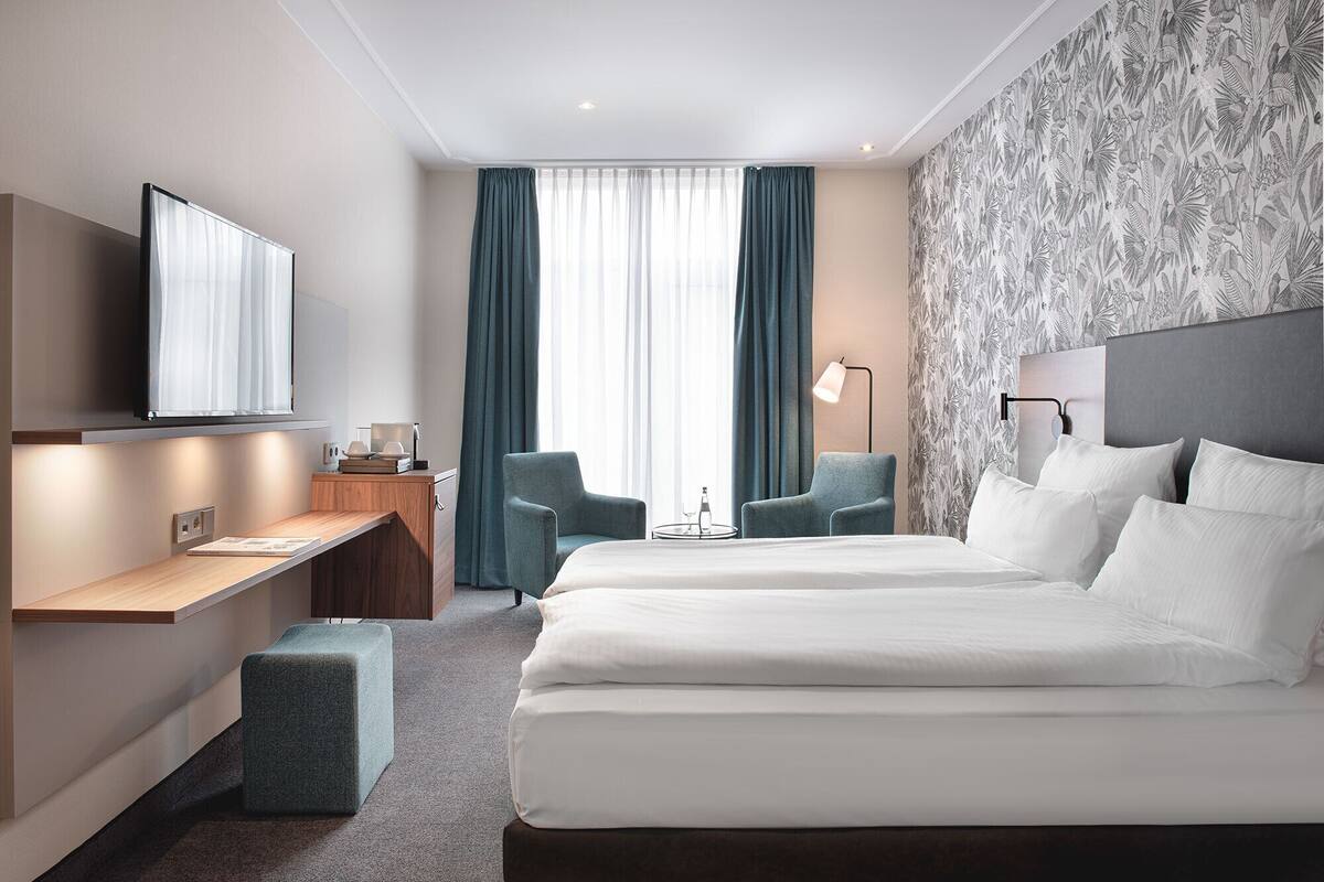 A hotel room with a bed, a desk, a chair, a TV, and a window with curtains.