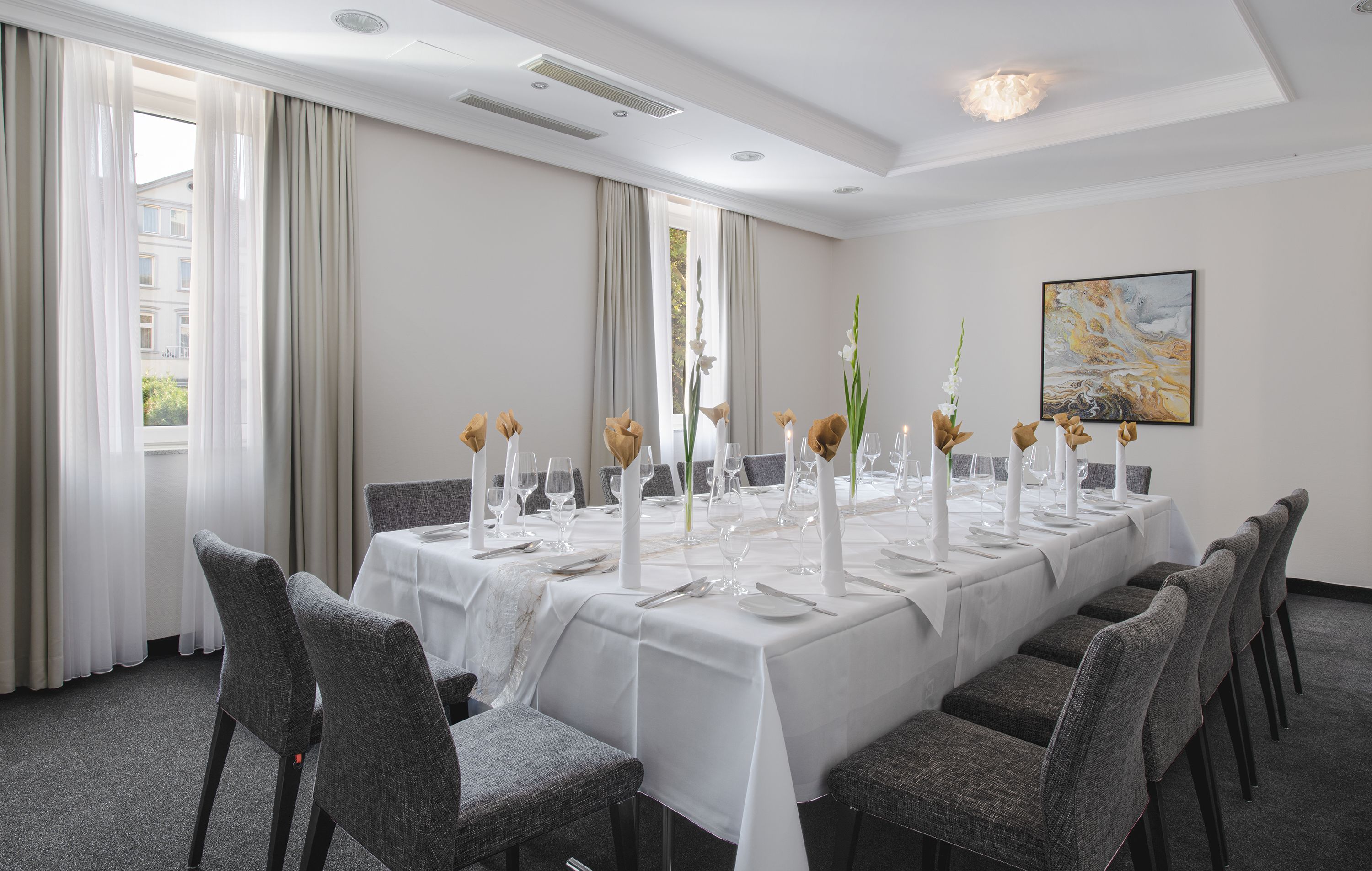 Una larga mesa de comedor cubierta con manteles blancos, velas y centros de mesa florales en una habitación bien iluminada con ventanas grandes y un candelabro.