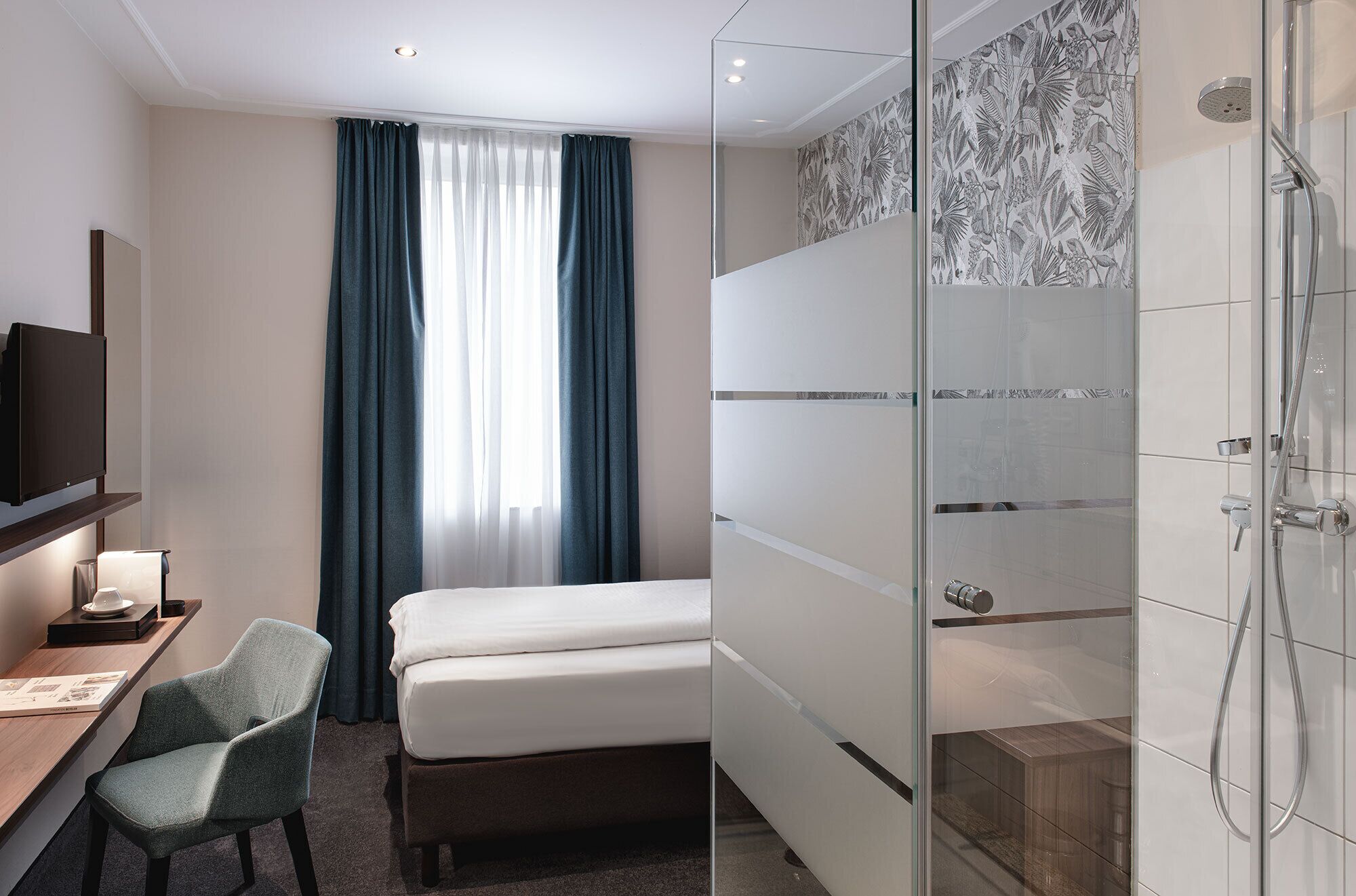 Ein Hotelzimmer mit Bett, Schreibtisch mit Stuhl, Fernseher und einer Dusche mit Glastür.