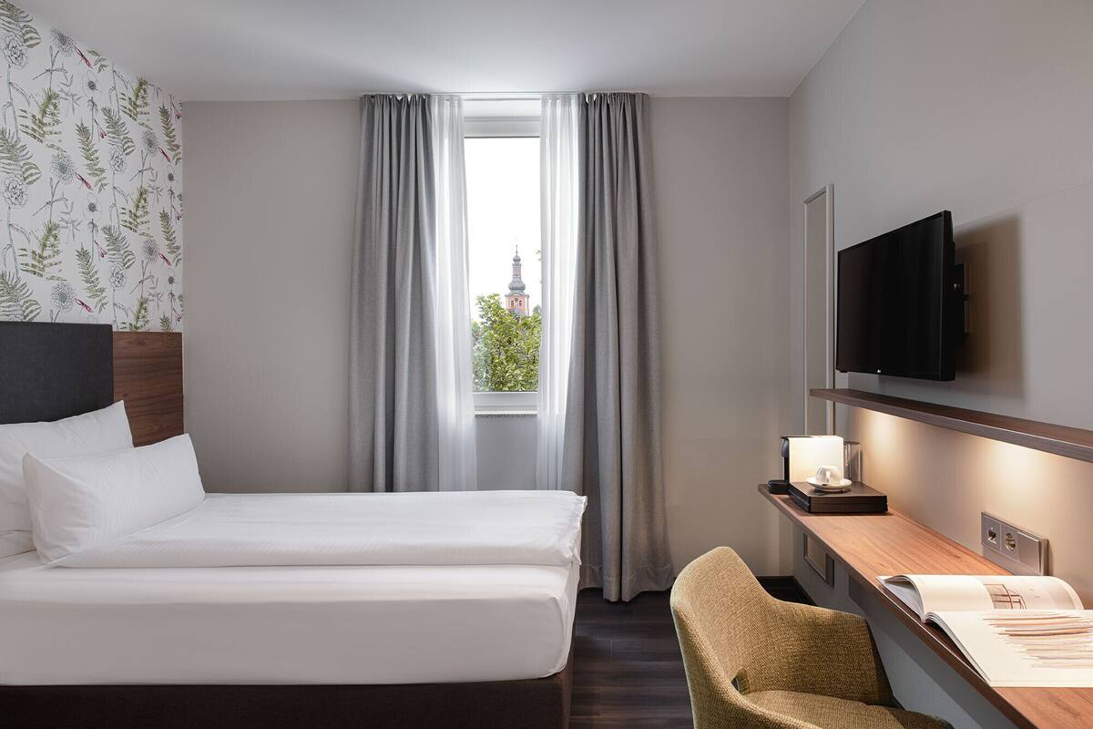 A hotel room with a bed, a desk with a lamp, a chair, and a TV.