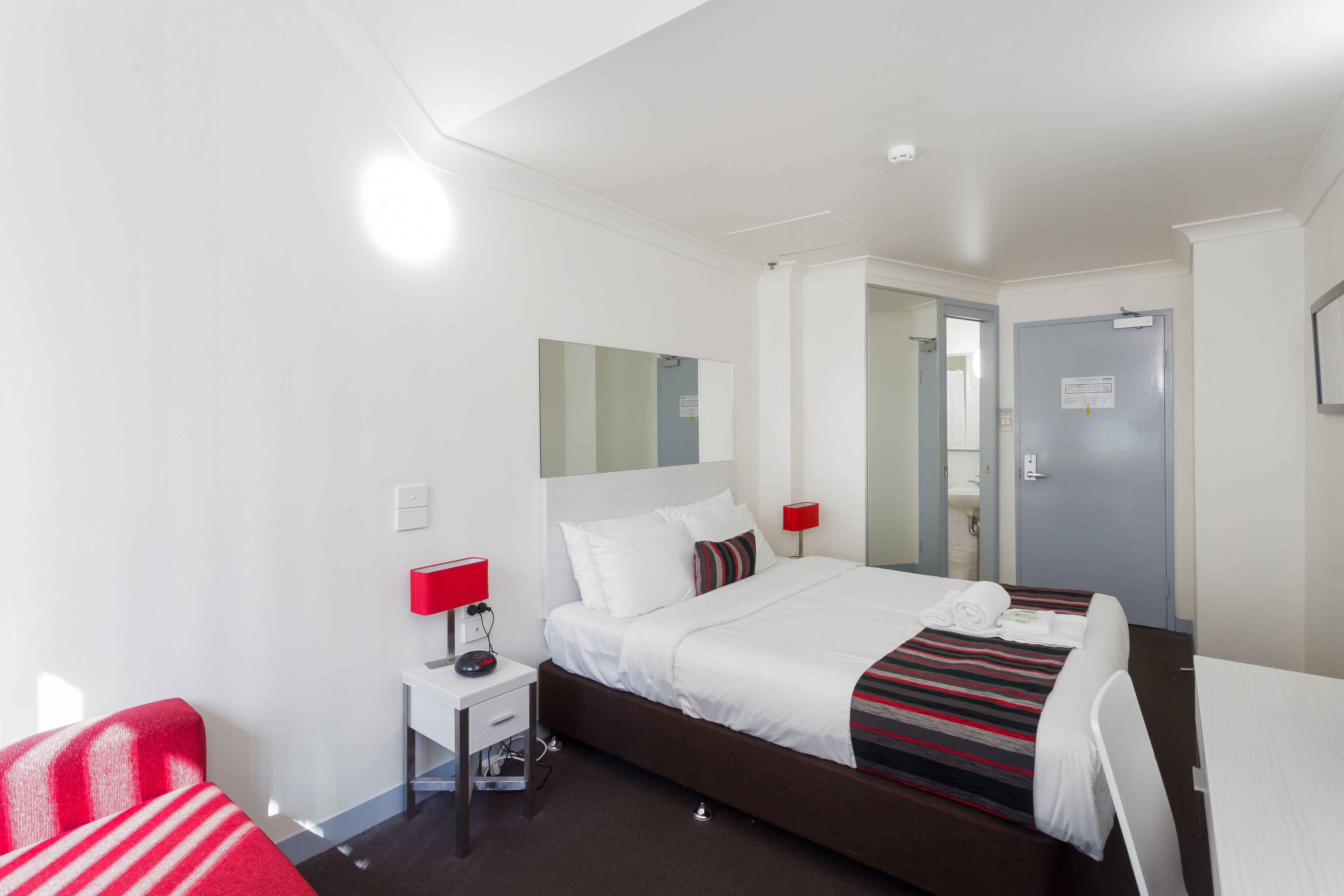 A hotel room with a large bed, a bedside table with a red lamp, a small desk, and a chair.