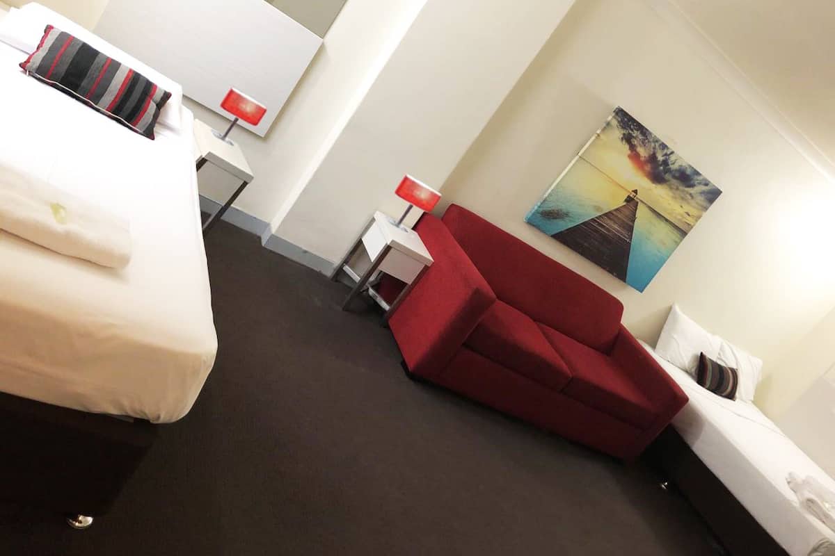 A hotel room with two beds, a red sofa, a small table with a lamp, and a painting on the wall.