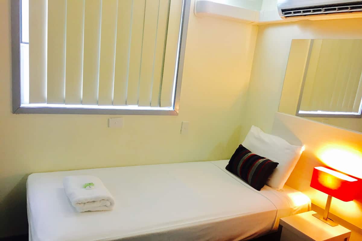 A small, neatly made bed with a white sheet and a folded towel, a bedside lamp, a window with blinds, and a wall-mounted air conditioning unit.
