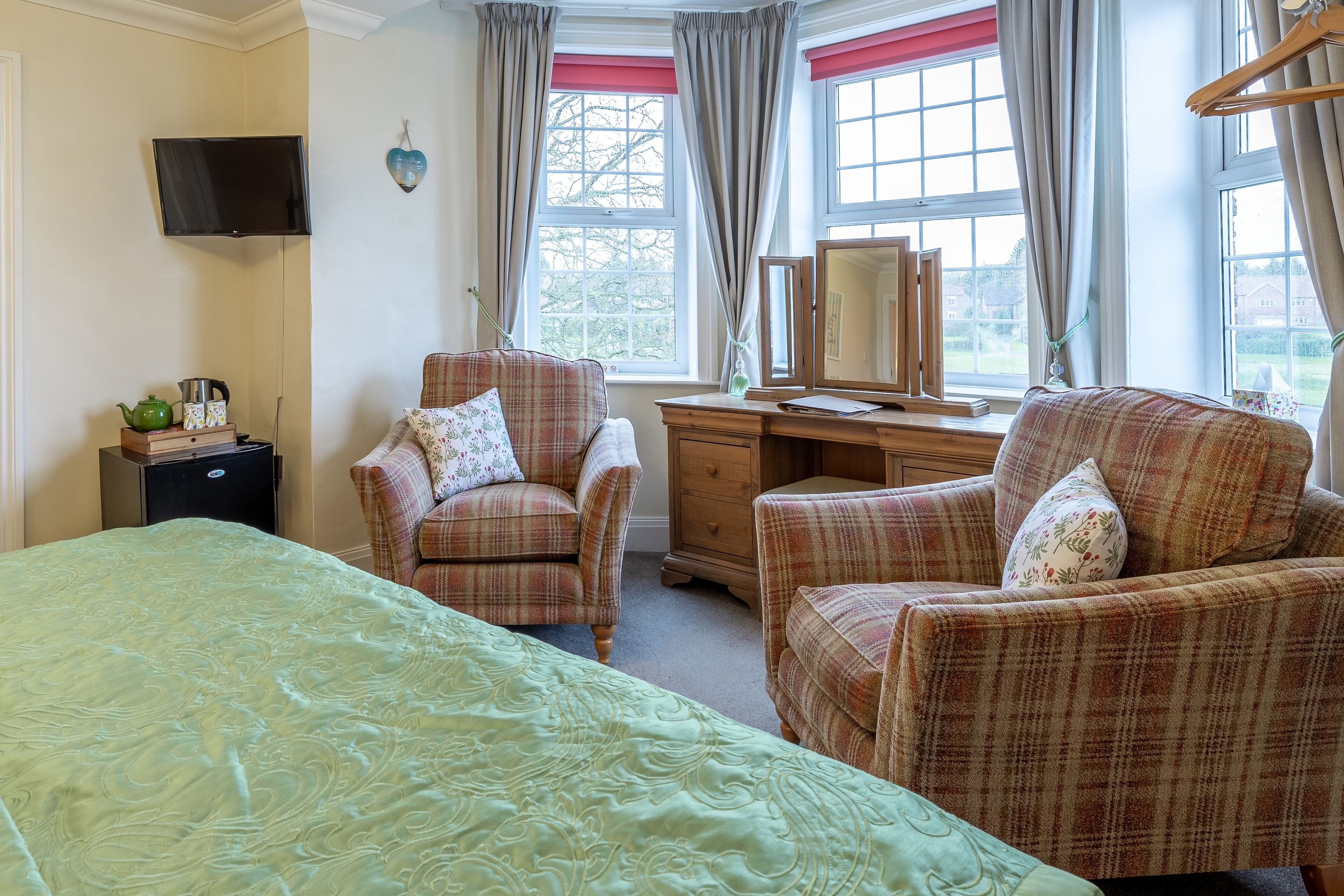 A bedroom with a bed, two armchairs, a dresser, a mirror, a television, and a window with curtains.