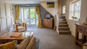 A cozy living room with a sofa, a wooden coffee table, a TV on a cabinet, a staircase, and a view of the outdoors through a window. - Harmony House (York)