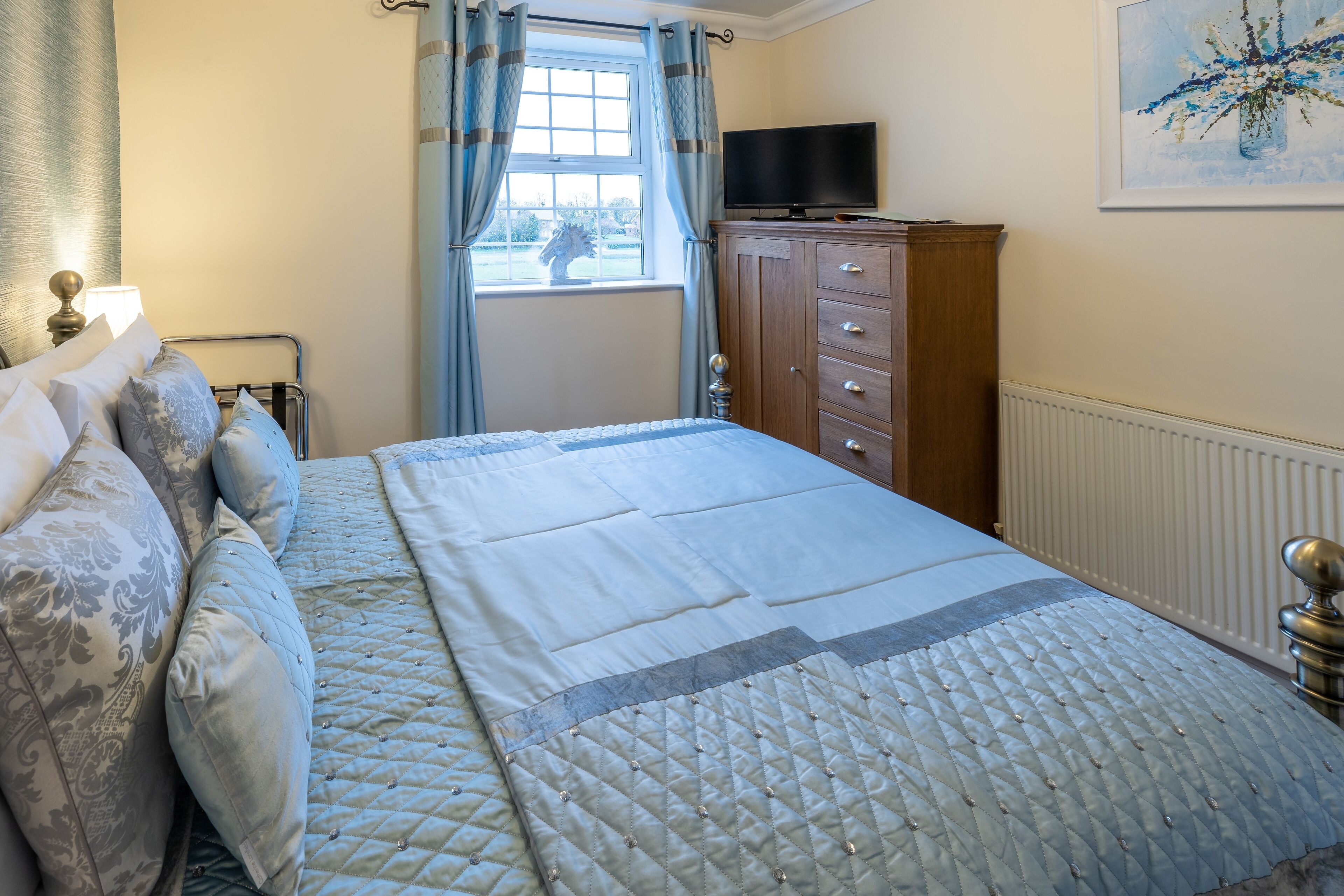 A neatly made bed with a quilted duvet, a wooden dresser with a flat-screen TV, and a window with curtains.