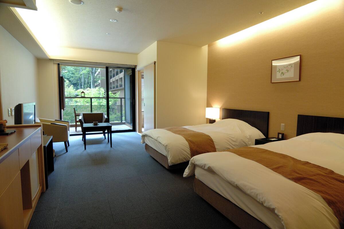 A hotel room with two beds, a desk, a chair, and a large window offering a view of greenery.