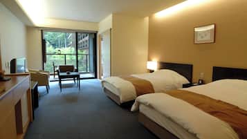 A hotel room with two beds, a desk, a chair, and a large window offering a view of greenery.
