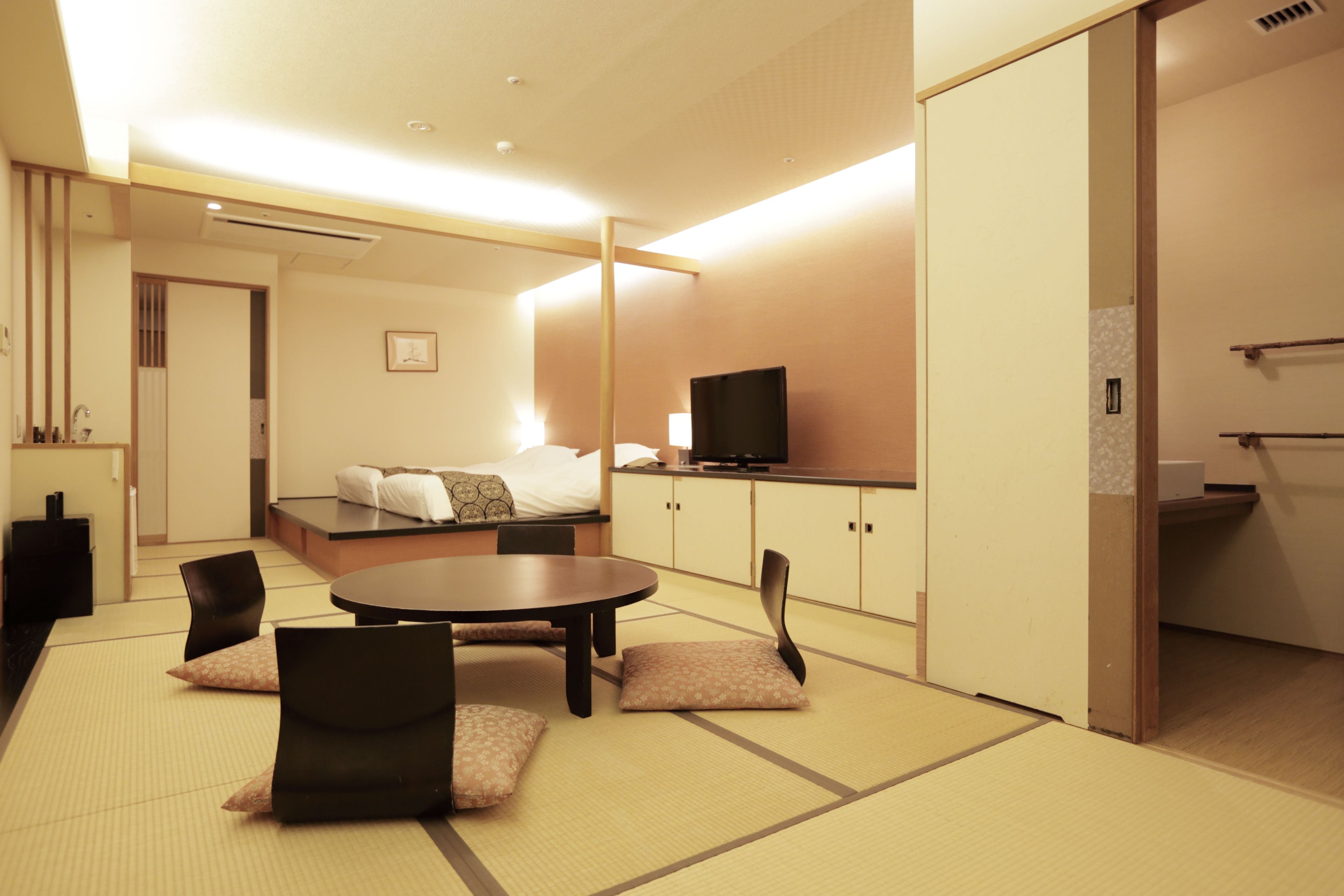 A modern hotel room with a bed, a TV, a dining area with tatami mats, and a kitchenette.