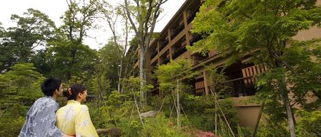 A traditional Japanese garden with a wooden bridge, lush greenery, and a building with multiple balconies.