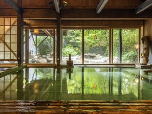 Un onsen japonés tradicional con interiores de madera, un amplio ventanal con vistas a un paisaje natural y una cuenca de piedra en el centro.
