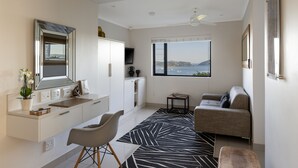 A modern living room with a sofa, a dining area with a white cabinet and a small table, a large window with a view of the sea, and a ceiling fan. - THEBLOEM Guest Suites by Knysna Paradise Collection (Knysna)