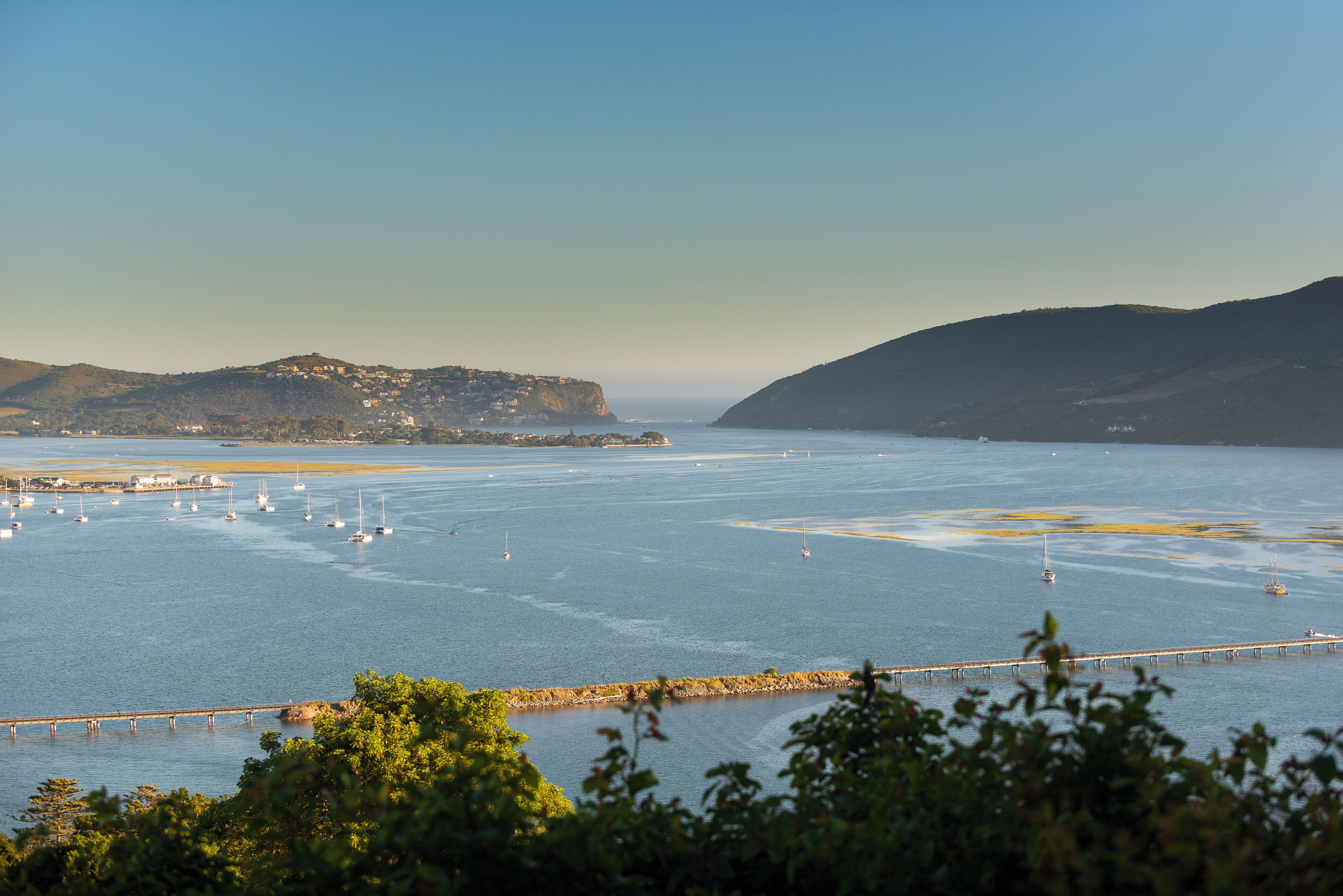 Vista costera con un muelle que se adentra en el agua, rodeado de colinas y un cielo despejado.