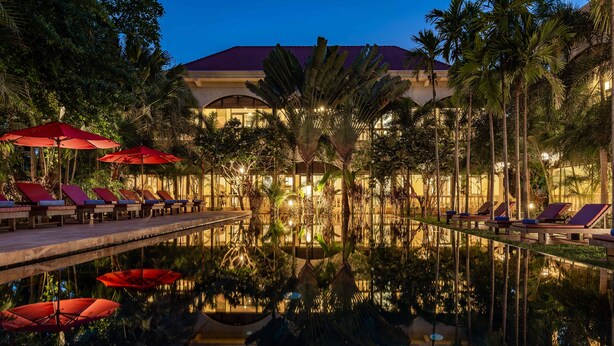 A pool area with lounge chairs and umbrellas, surrounded by palm trees and a building with a red roof. - Pavillon d'Orient Boutique (Siem Reap)