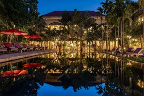 A pool area with lounge chairs and umbrellas, surrounded by palm trees and a building with a red roof. - Pavillon d'Orient Boutique (Siem Reap)