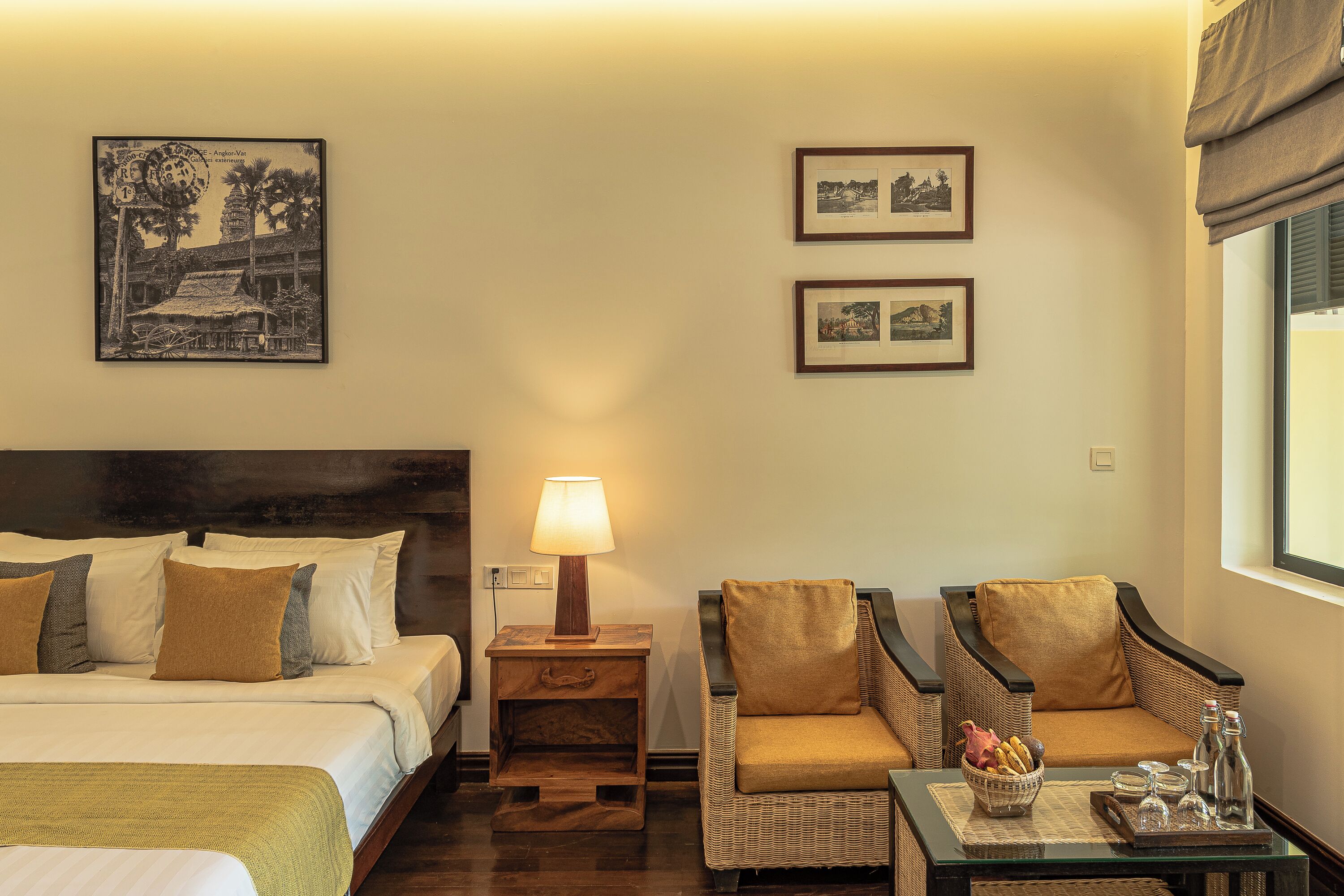 Camera d'albergo con un letto, due poltrone, un comodino con una lampada, un tavolino con un cesto di pane e condimenti, e due quadri appesi al muro.