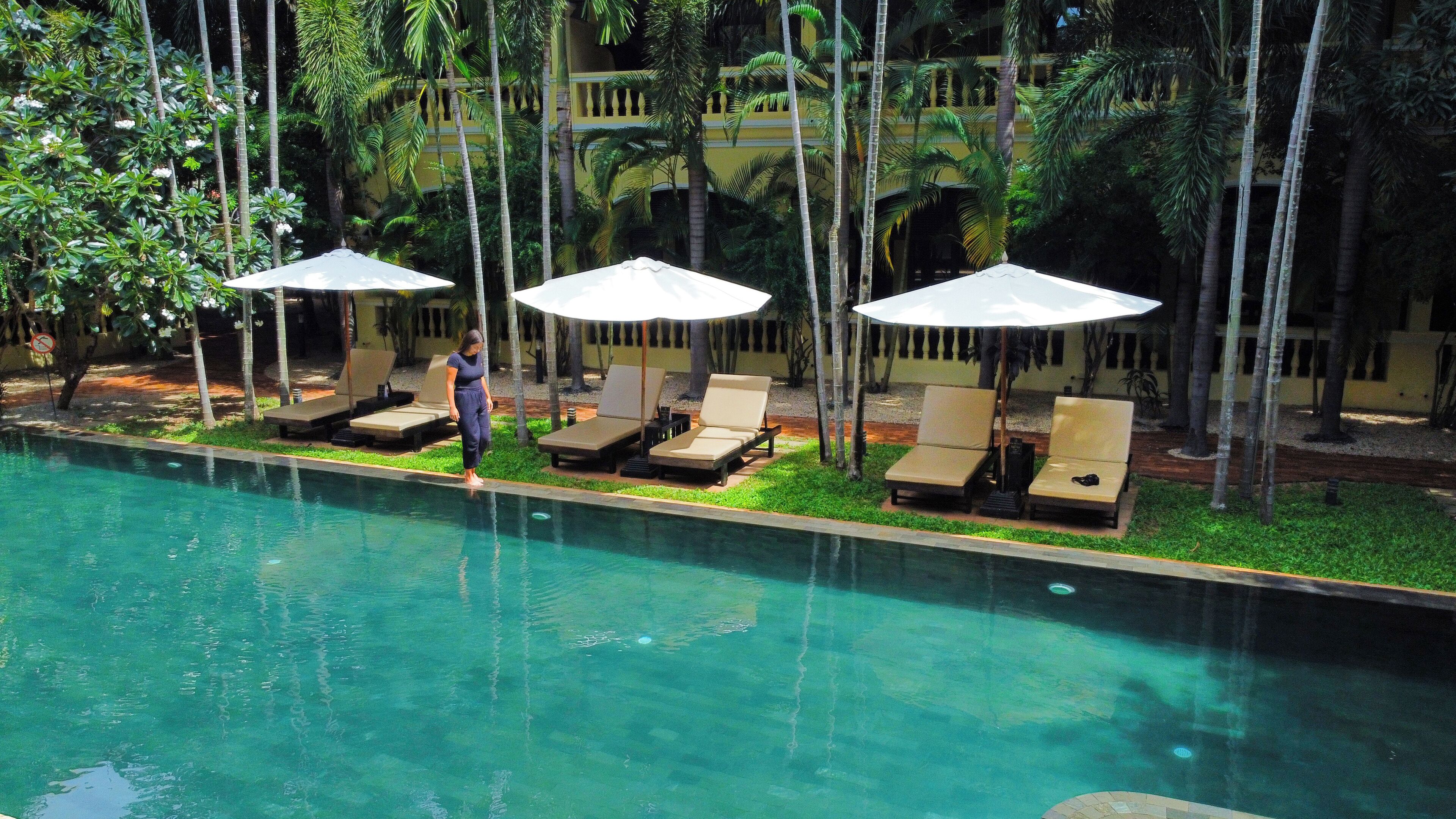 Zona junto a la piscina con sillones de descanso y sombrillas, rodeada de palmeras y un edificio al fondo.
