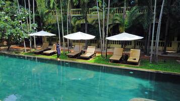 Zona junto a la piscina con sillones de descanso y sombrillas, rodeada de palmeras y un edificio al fondo.